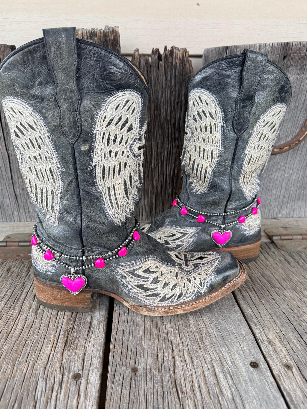 Pair of black angel wing embroidered cowboy boots wearing double-strand silver and hot pink beaded boot bracelets with pink enamel heart charms, set of 2