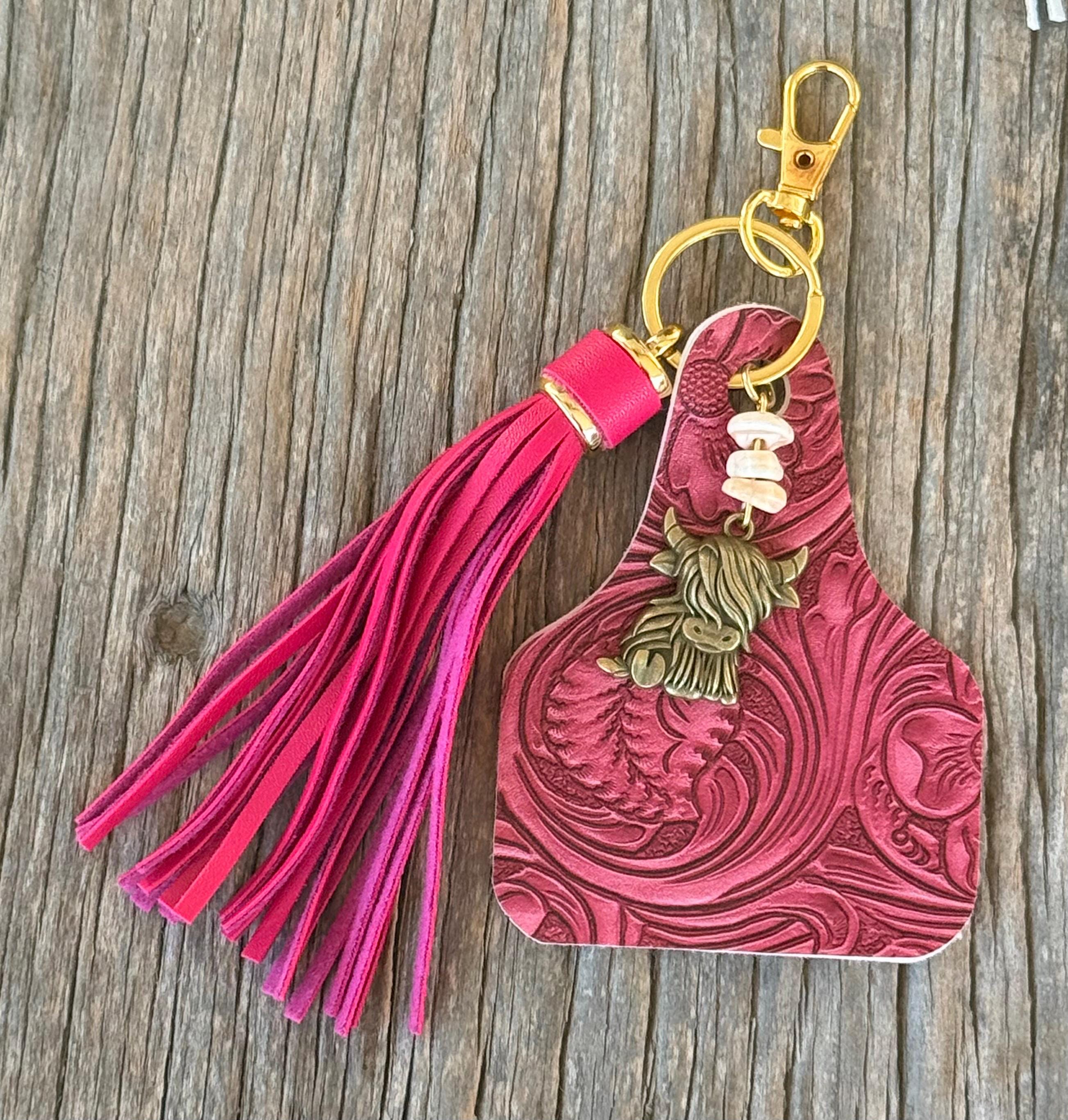 Flat lay of hot pink tooled leather cow tag keychain bag charm with pink leather fringe tassel, white shell bead stack, and bronze highland cow charm on gold lobster clasp and ring, on weathered wood planks