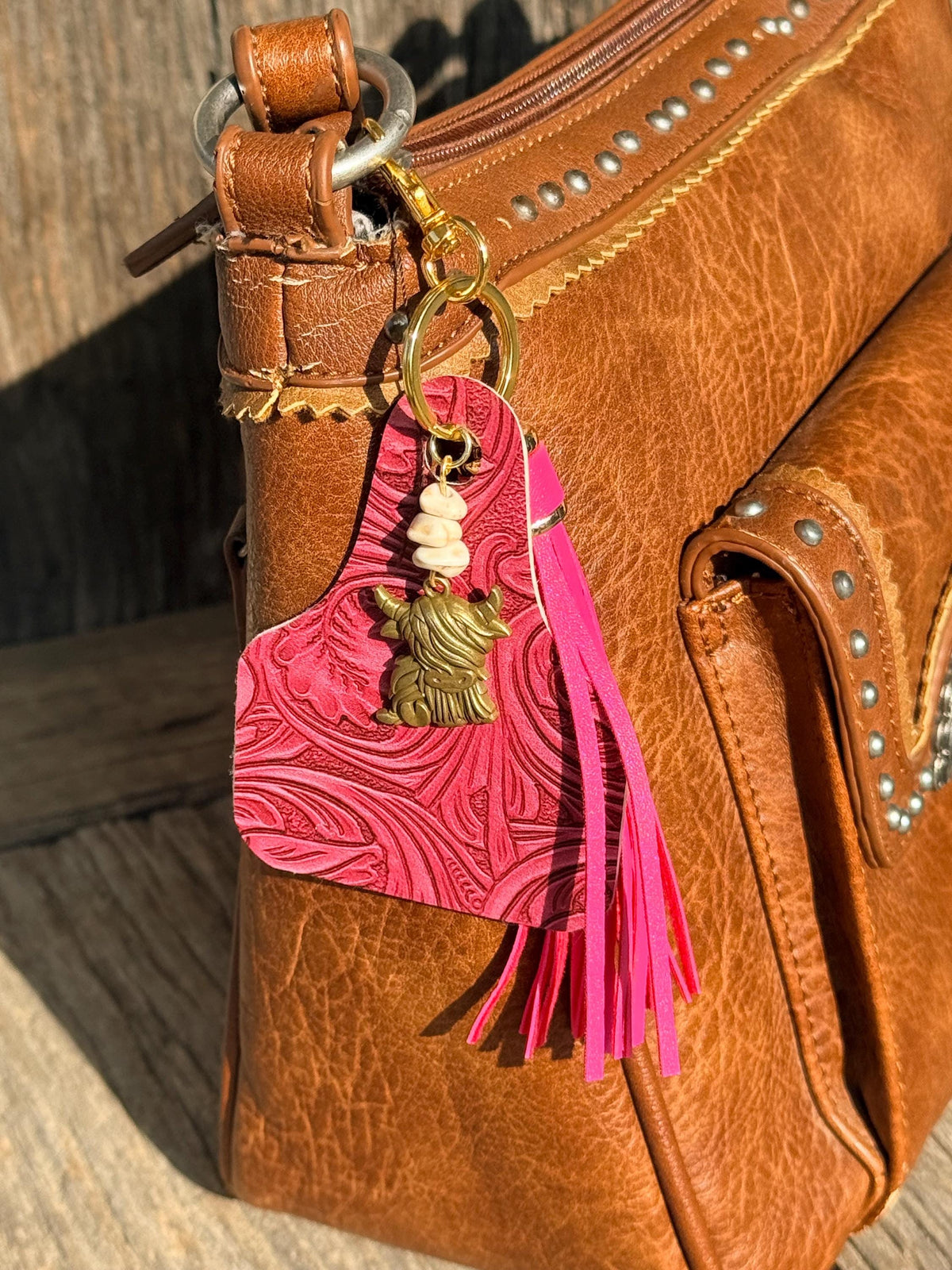 Hot pink tooled leather cow tag keychain bag charm with pink leather fringe tassel, white shell bead stack, and bronze highland cow charm clipped to brown studded leather handbag on weathered wood, Western boho purse accessory
