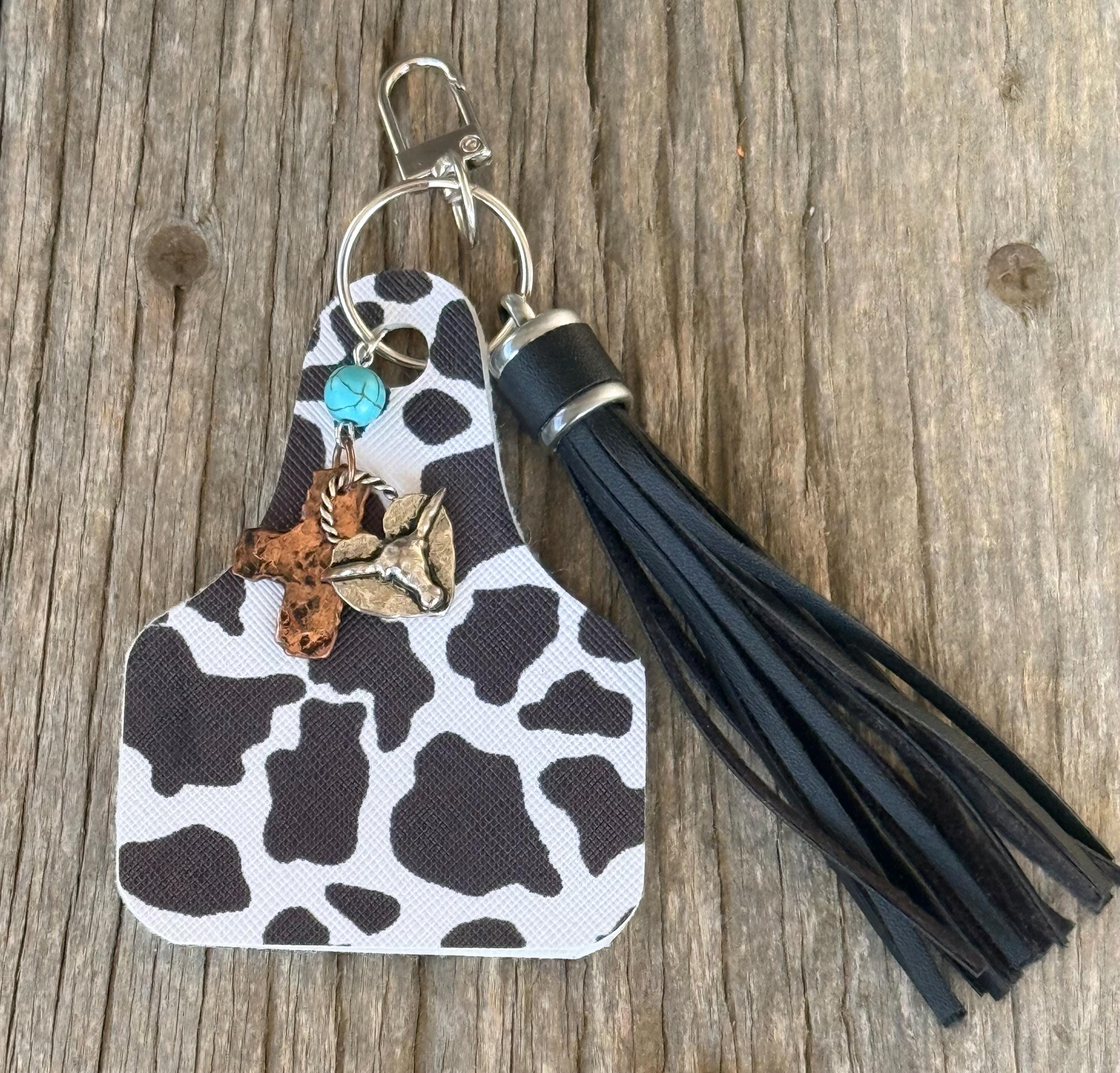 Flat lay of black and white cow print cow tag keychain bag charm with black leather fringe tassel, turquoise bead, bronze longhorn skull, and gold arrow cross charm on silver lobster clasp and ring, on weathered wood planks