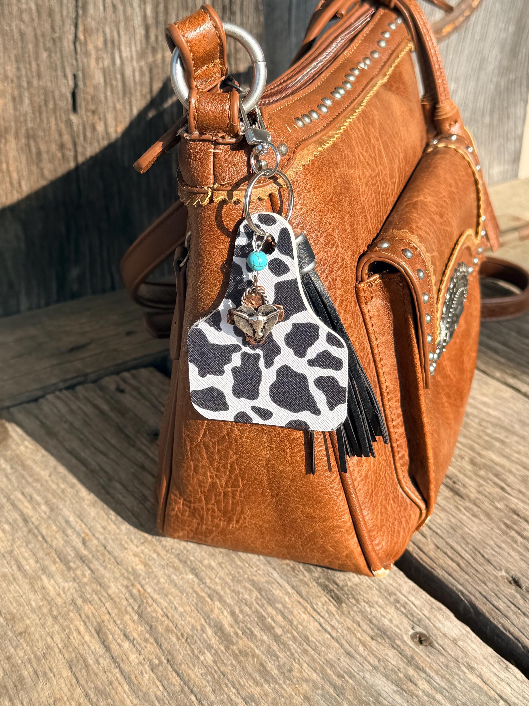 Black and white cow print cow tag keychain bag charm with black leather fringe tassel, turquoise bead, bronze longhorn skull, and gold arrow cross charm clipped to brown studded leather handbag on weathered wood, Western boho purse accessory