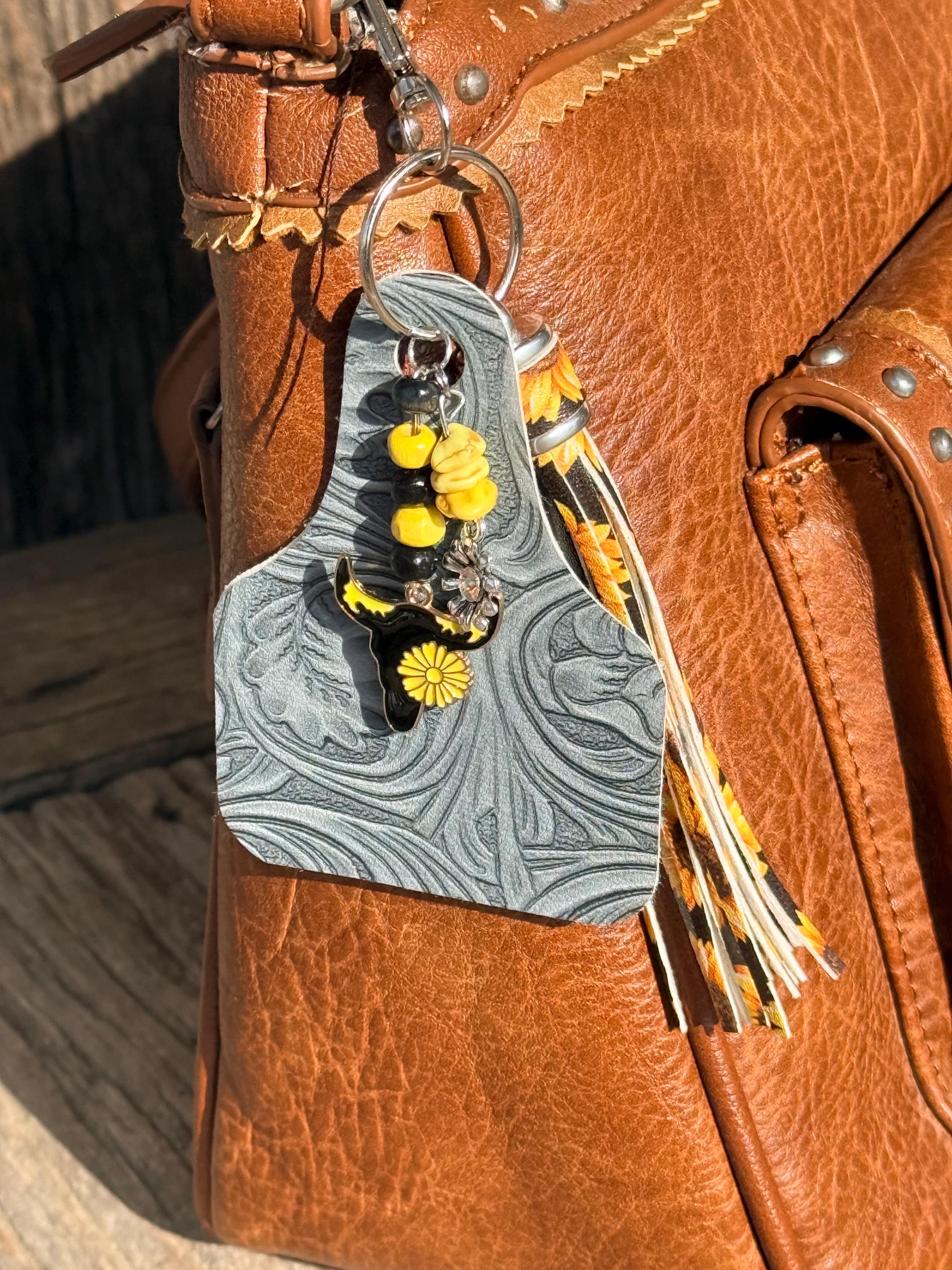 Gray-blue tooled leather cow tag keychain bag charm with sunflower print fabric fringe tassel, yellow turquoise chip beads, and black enamel longhorn skull with yellow sunflower charm clipped to brown studded leather handbag, Western boho purse accessory