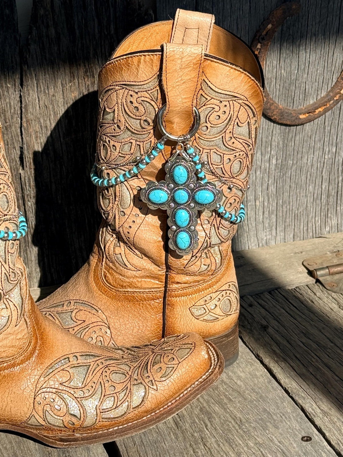 Angled front view of tan tooled leather cowboy boot with turquoise beaded boot bracelet and large silver turquoise cross concho charm, rustic Western setting