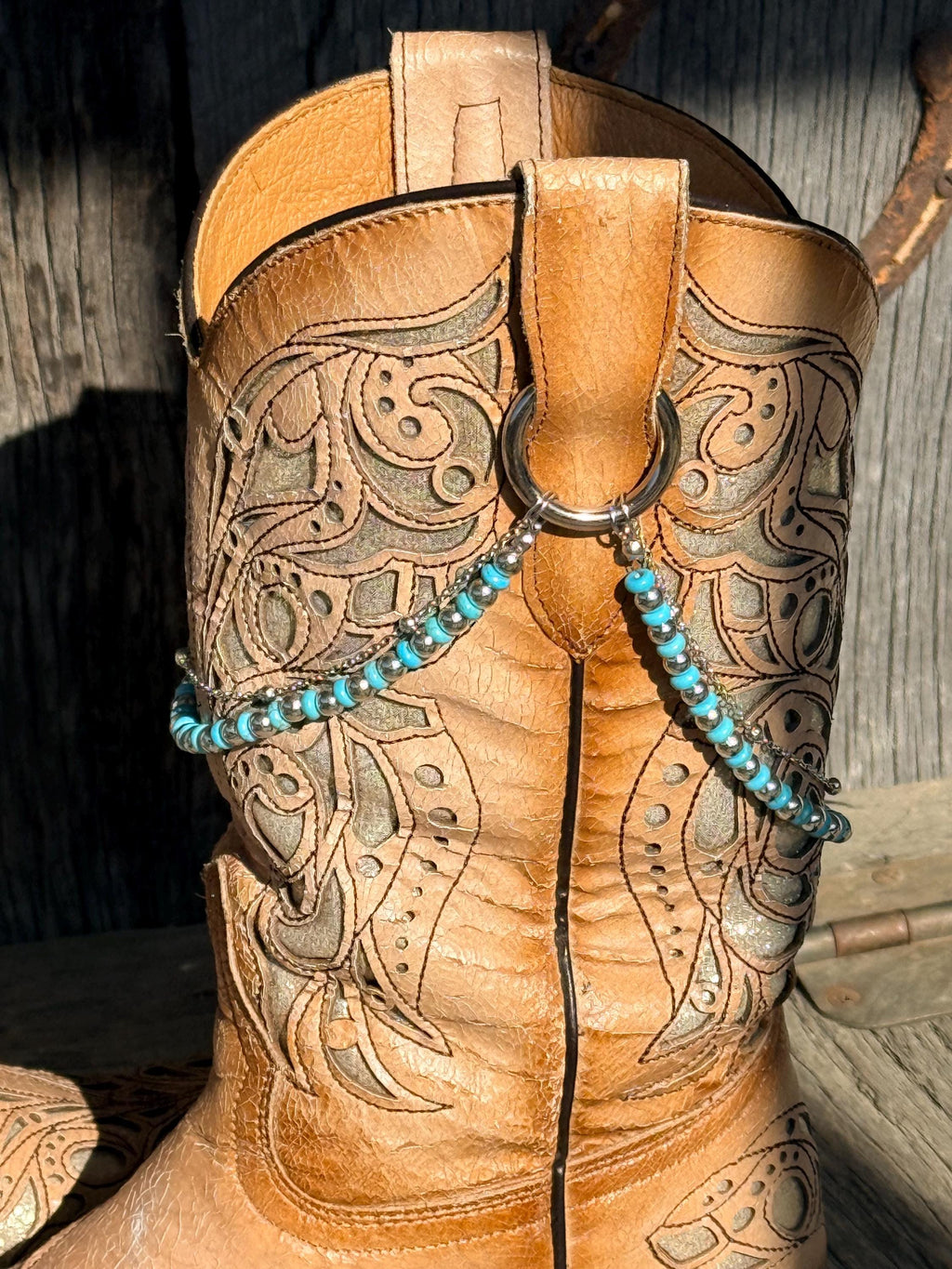A close-up of a tan leather western boot with intricate tooling and a silver-toned ring closure, decorated with a turquoise beaded chain.