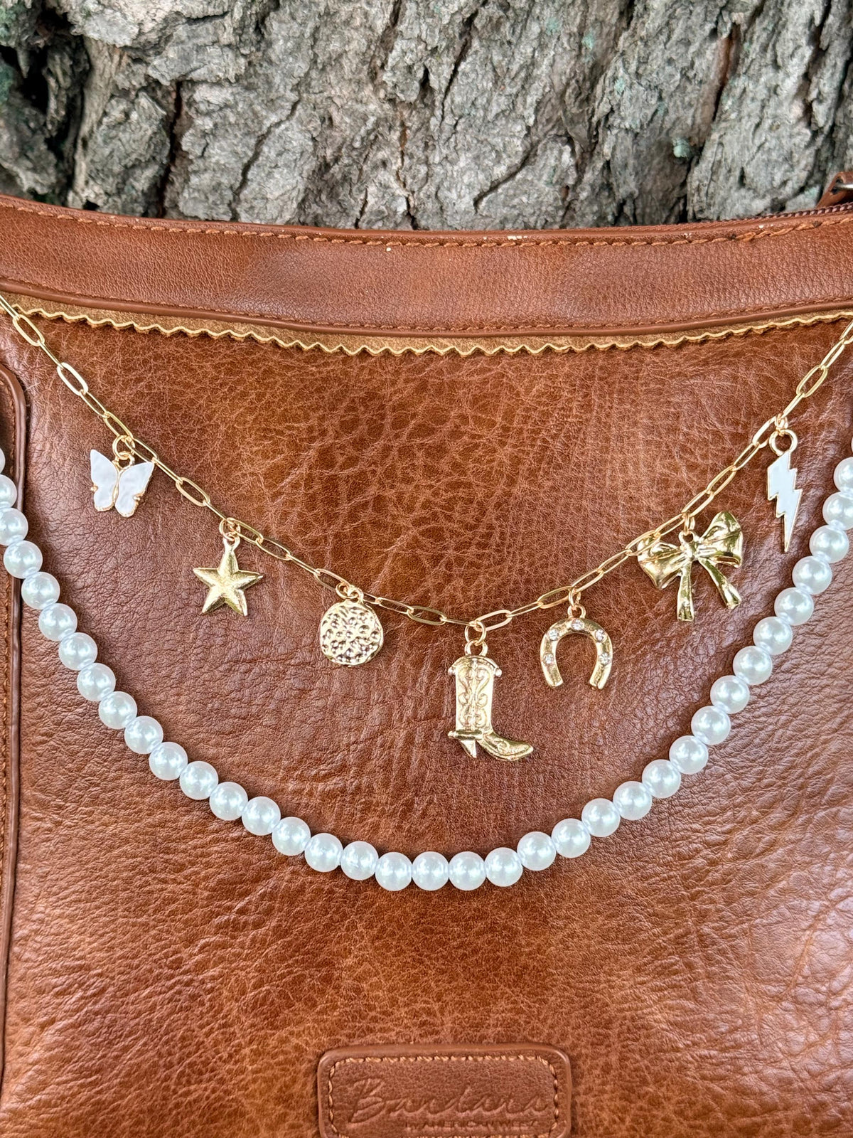 Close-up of brown distressed Bandana leather crossbody bag with gold paperclip link chain featuring seven Western charms: white enamel butterfly, gold star, gold coin medallion, gold cowboy boot, gold horseshoe, gold bow, and gold lightning bolt, with faux pearl bead strand of uniform round pearls draping below, Western boho 11-inch purse chain detail