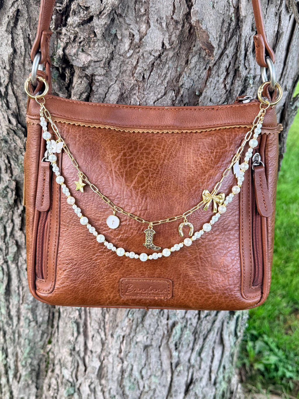 Front view of brown distressed Bandana leather crossbody bag against tree with two-strand Western boho purse chain featuring gold paperclip link chain with six Western charms above and mixed-size pearl bead strand with gold rondelle spacers draping below, 13-inch Western handbag jewelry