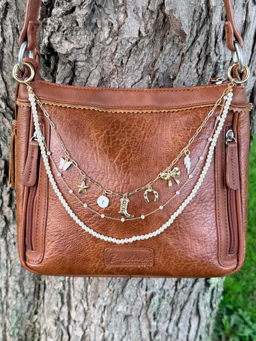 Front view of brown leather Bandana crossbody bag against tree with three-strand Western boho purse chain featuring gold paperclip link chain with seven Western charms including white butterfly, gold star, cross coin disc, ornate cowboy boot, horseshoe, bow, and lightning bolt, plus delicate gold chain with pink bead accents and faux pearl bead strand