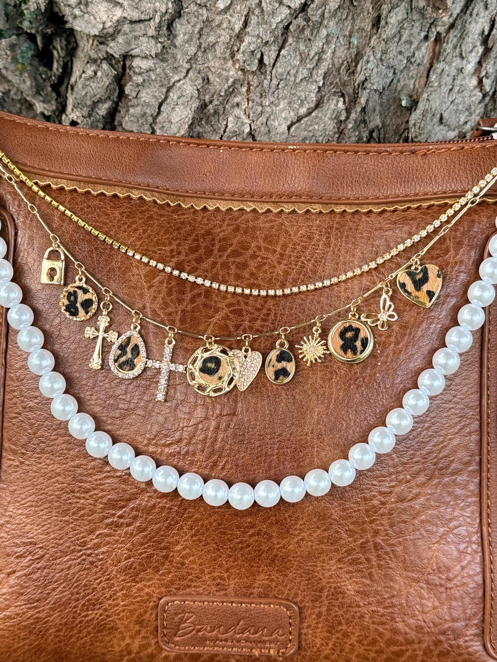 Close-up of brown distressed Bandana leather crossbody bag with gold paperclip link chain featuring eleven cheetah print and gold charms including crystal cross, gold padlock, gold sunburst, gold butterfly, and multiple cheetah print disc and heart charms, with gold crystal rhinestone tennis chain and faux pearl bead strand, Western boho 10-inch purse chain detail