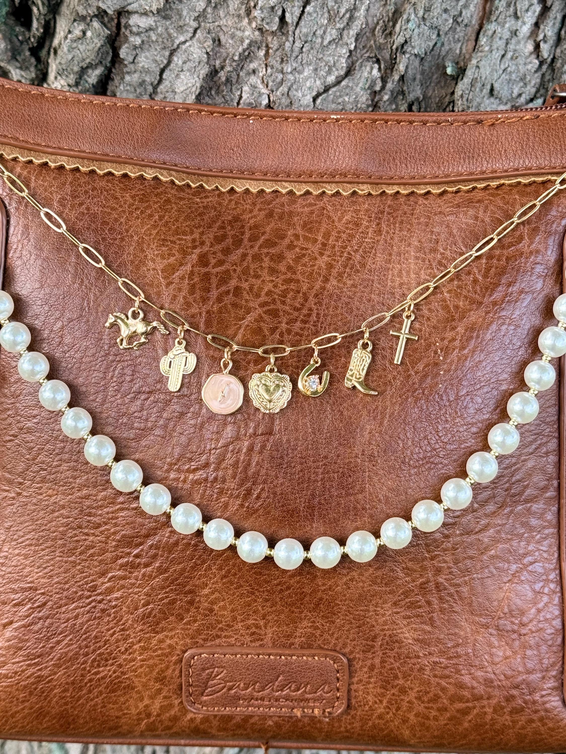 Close-up of brown leather Bandana crossbody bag with gold paperclip link chain featuring seven Western charms including horse and rider, cactus, pink enamel coin, ornate gold heart, crystal cowgirl boot, cowboy boot, and small cross, with faux pearl bead strand below