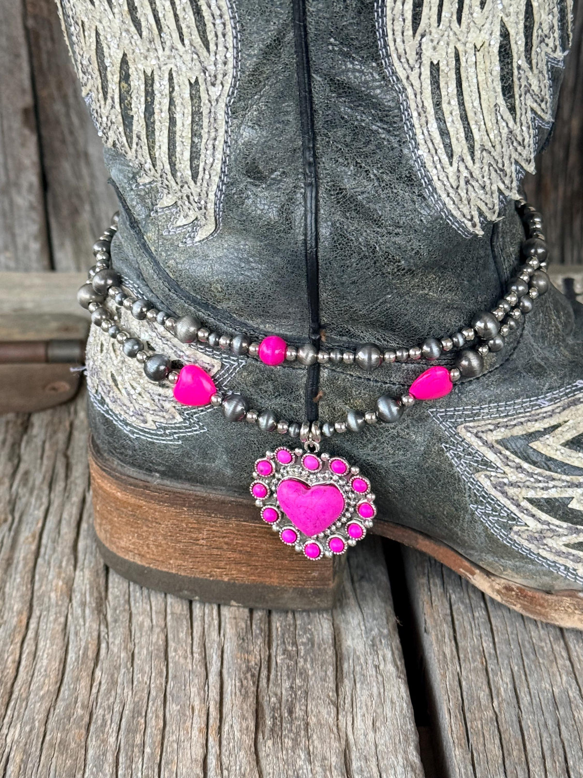 Side angle of black angel wing cowboy boot with double-strand silver and hot pink beaded boot bracelet and ornate pink rhinestone heart concho charm pendant