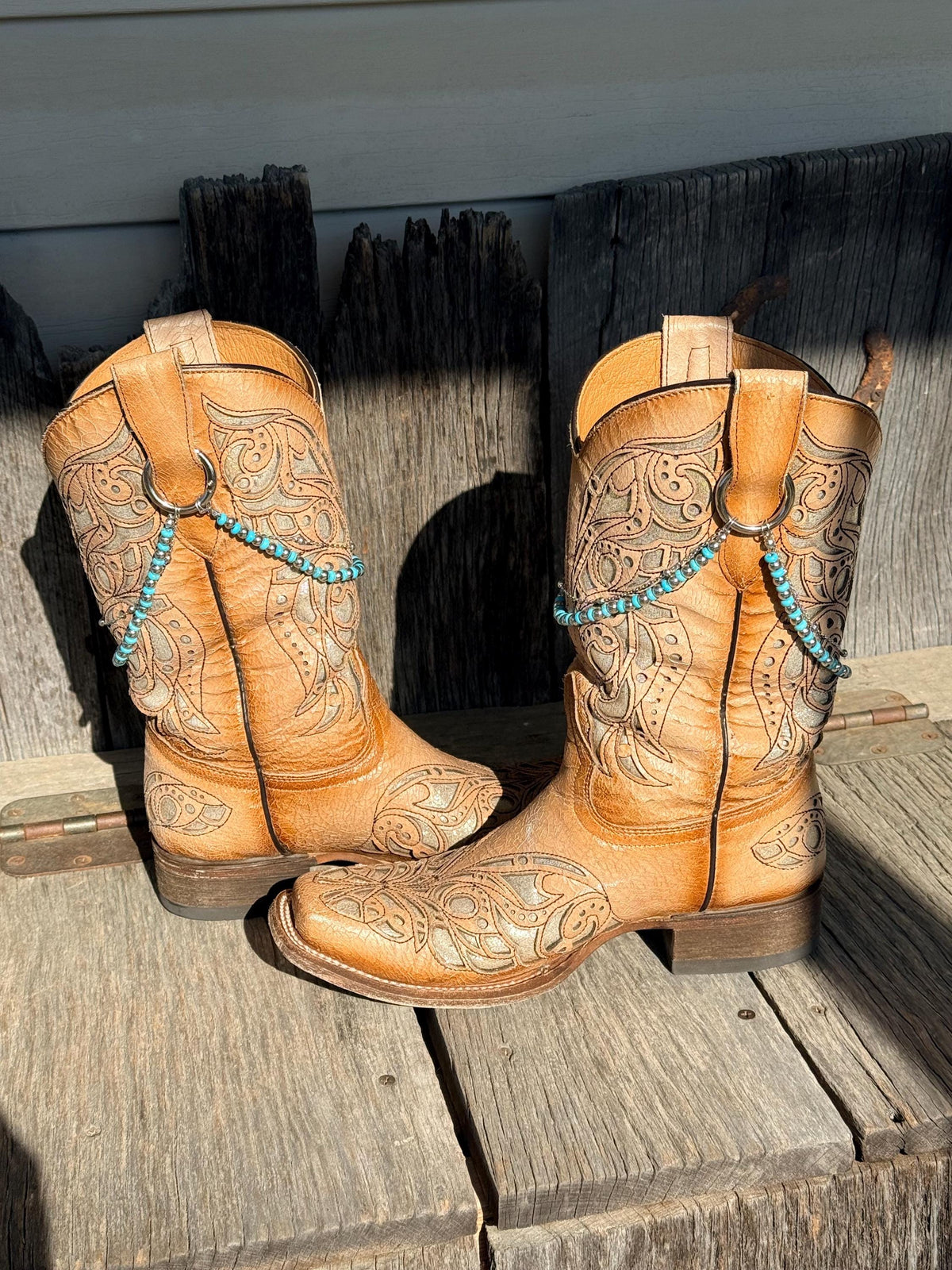 A pair of tan tooled leather cowboy boots with turquoise bead accents, displayed on rustic wooden planks.