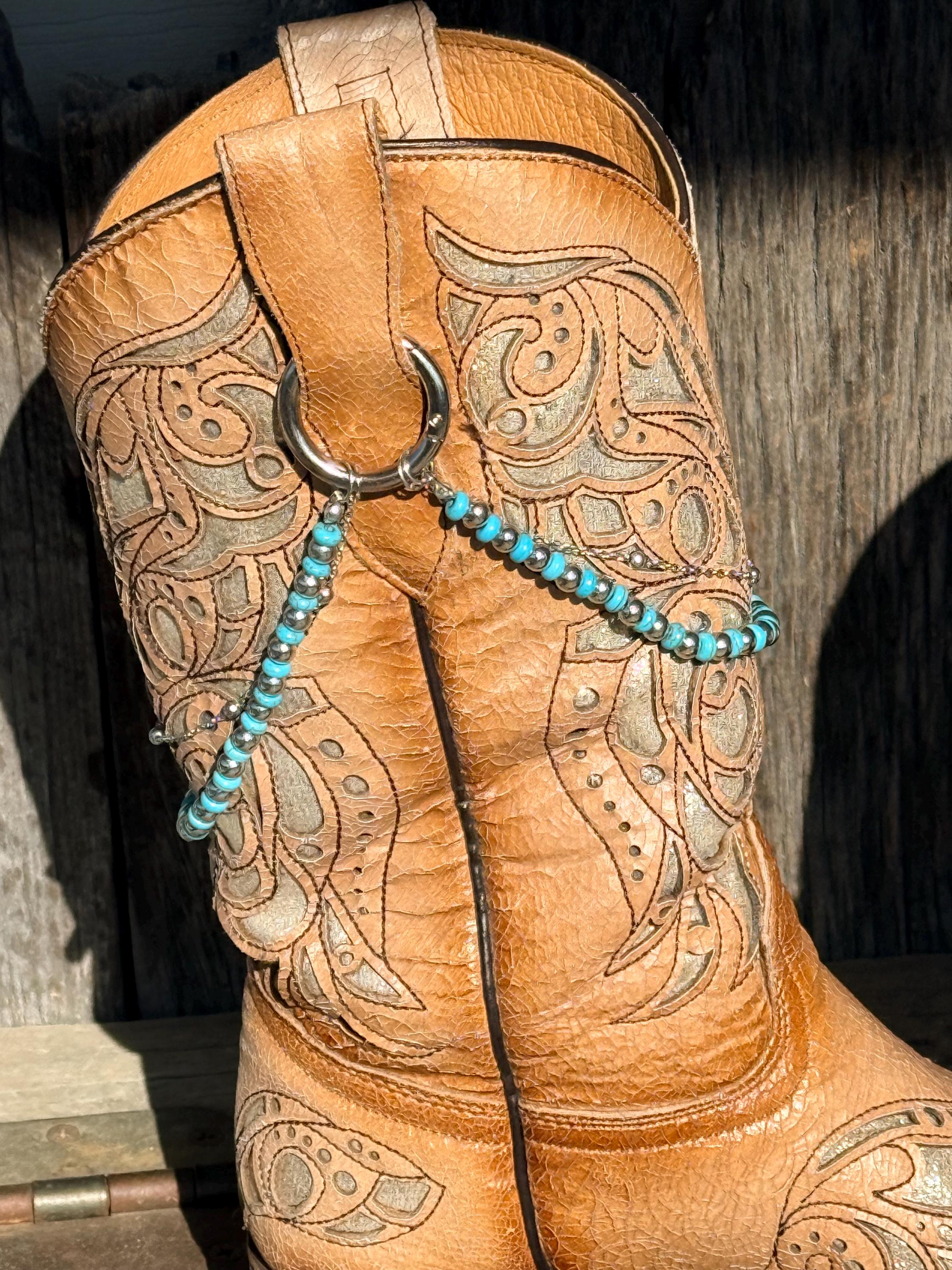 A close-up of a tan leather western boot with intricate tooling, featuring a turquoise beaded chain with a silver ring attached near the top.
