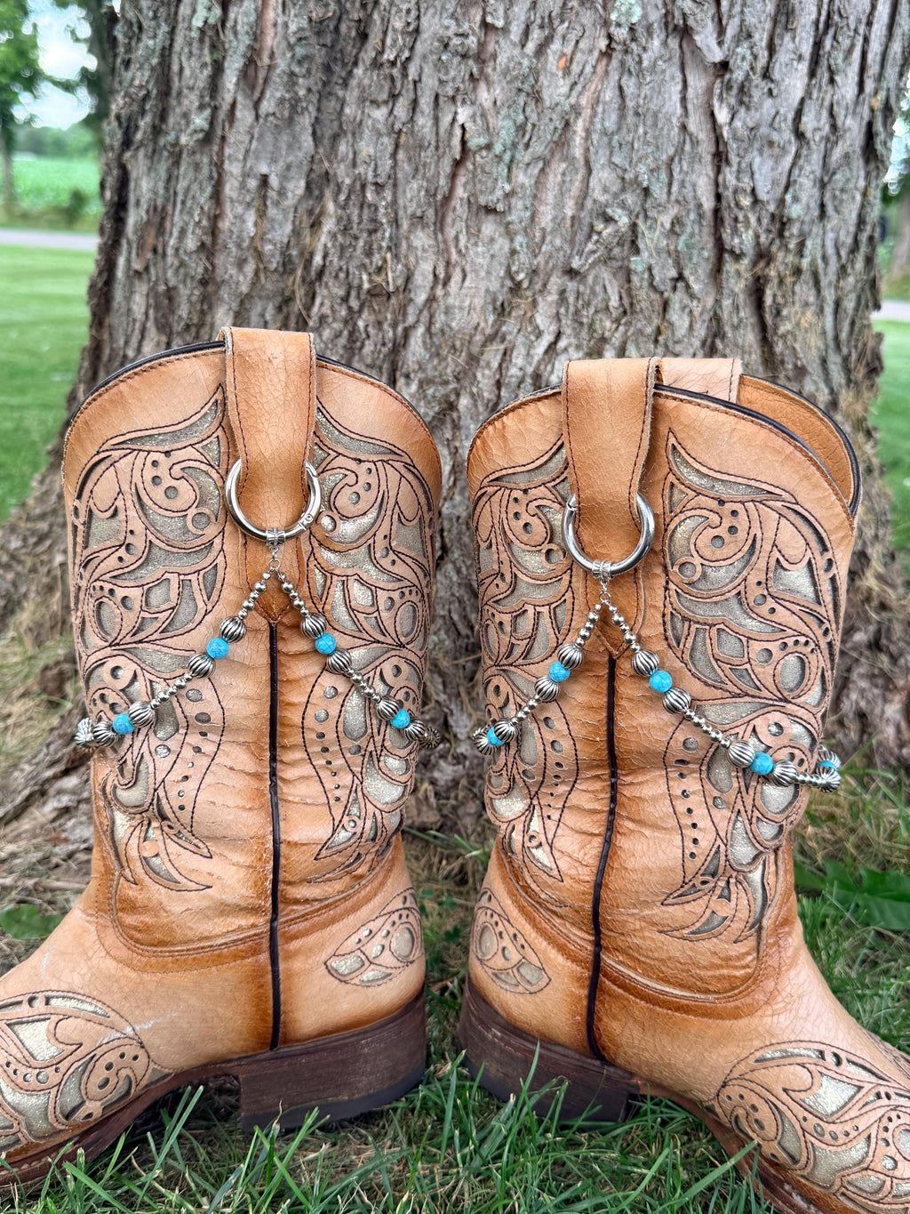 Back view of pair of tan laser-cut scrollwork cowboy boots on grass against tree showing silver ball chain and turquoise bead boot bracelet leis with silver ribbed spacers clipped through boot pull loop rings, Western boot bling set of 2