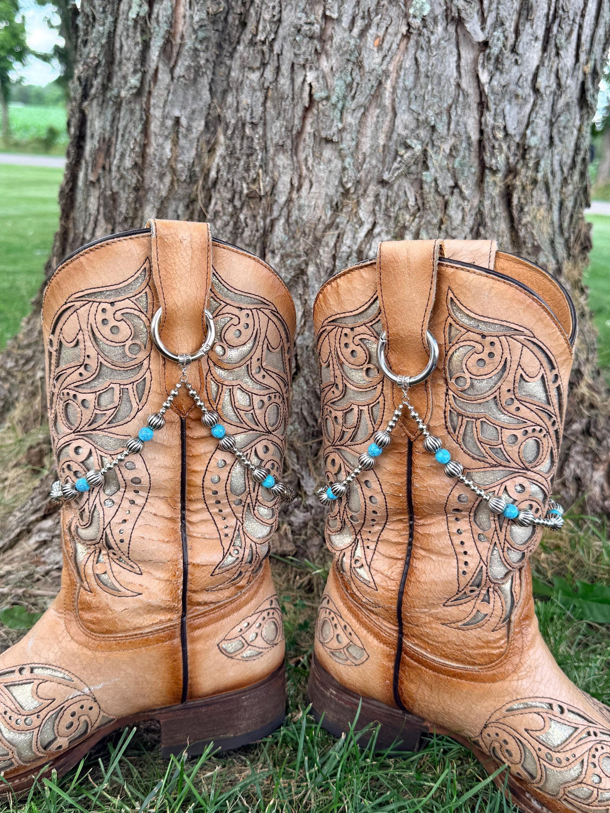 Back view of pair of tan laser-cut scrollwork cowboy boots on grass against tree showing silver ball chain and turquoise bead boot bracelet leis with silver ribbed spacers clipped through boot pull loop rings, Western boot bling set of 2