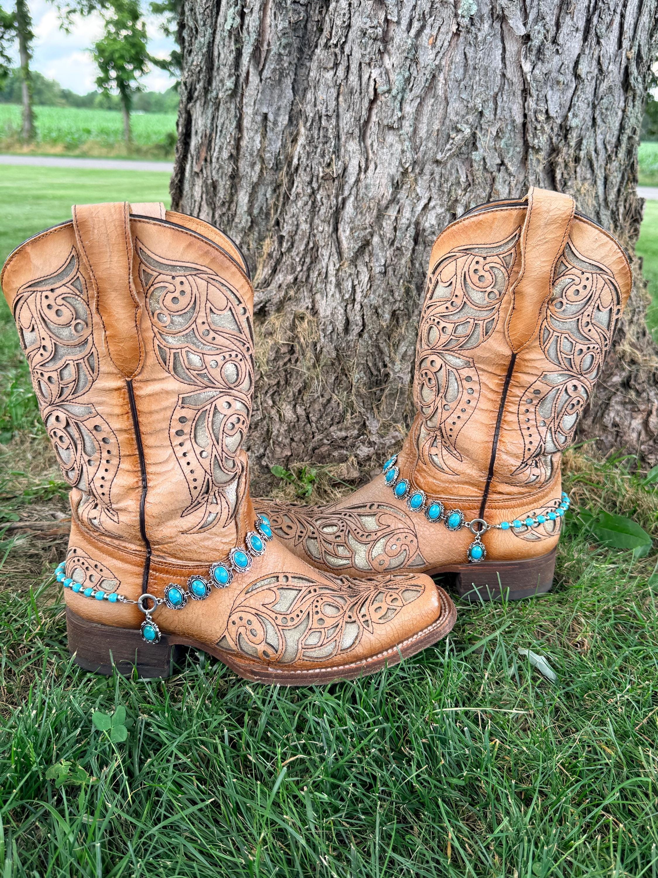 Front view of pair of tan tooled laser-cut cowboy boots against tree on grass wearing two-strand silver and turquoise boot bracelets with oval concho strand, bead strand, and turquoise drop charm, set of 2