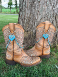 Front view of pair of tan tooled laser-cut cowboy boots against tree on grass wearing two-strand turquoise bead and silver chain lei boot bracelets with large turquoise stone heart and silver filigree overlay pendant, set of 2