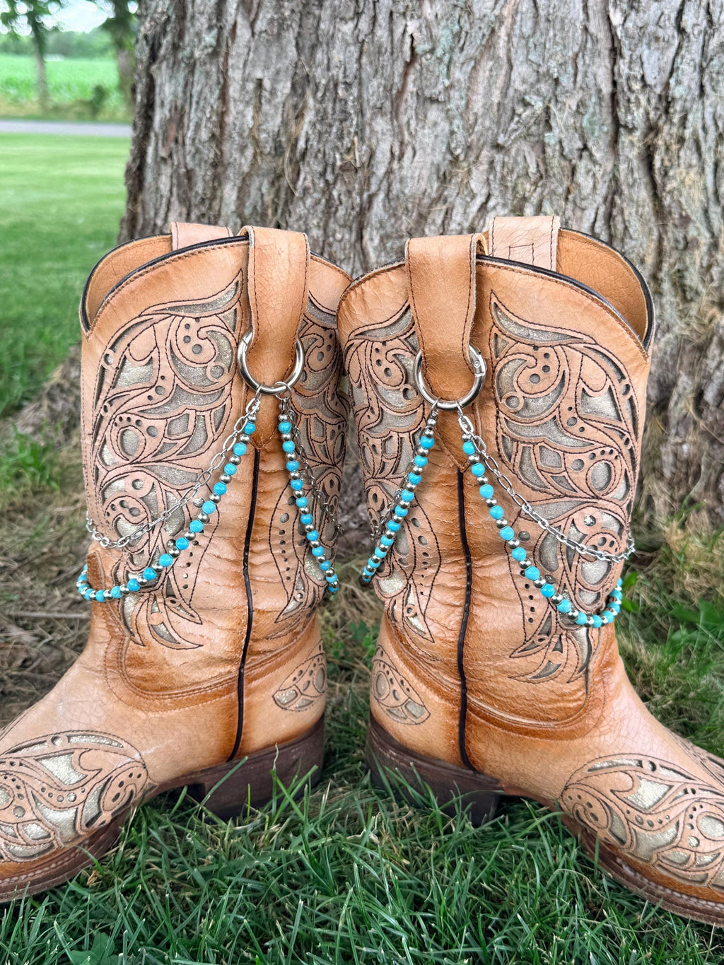 Back view of pair of tan tooled laser-cut cowboy boots against tree showing two-strand turquoise bead and silver chain lei boot bracelets clipped through boot pull loop rings, Western boho style set of 2