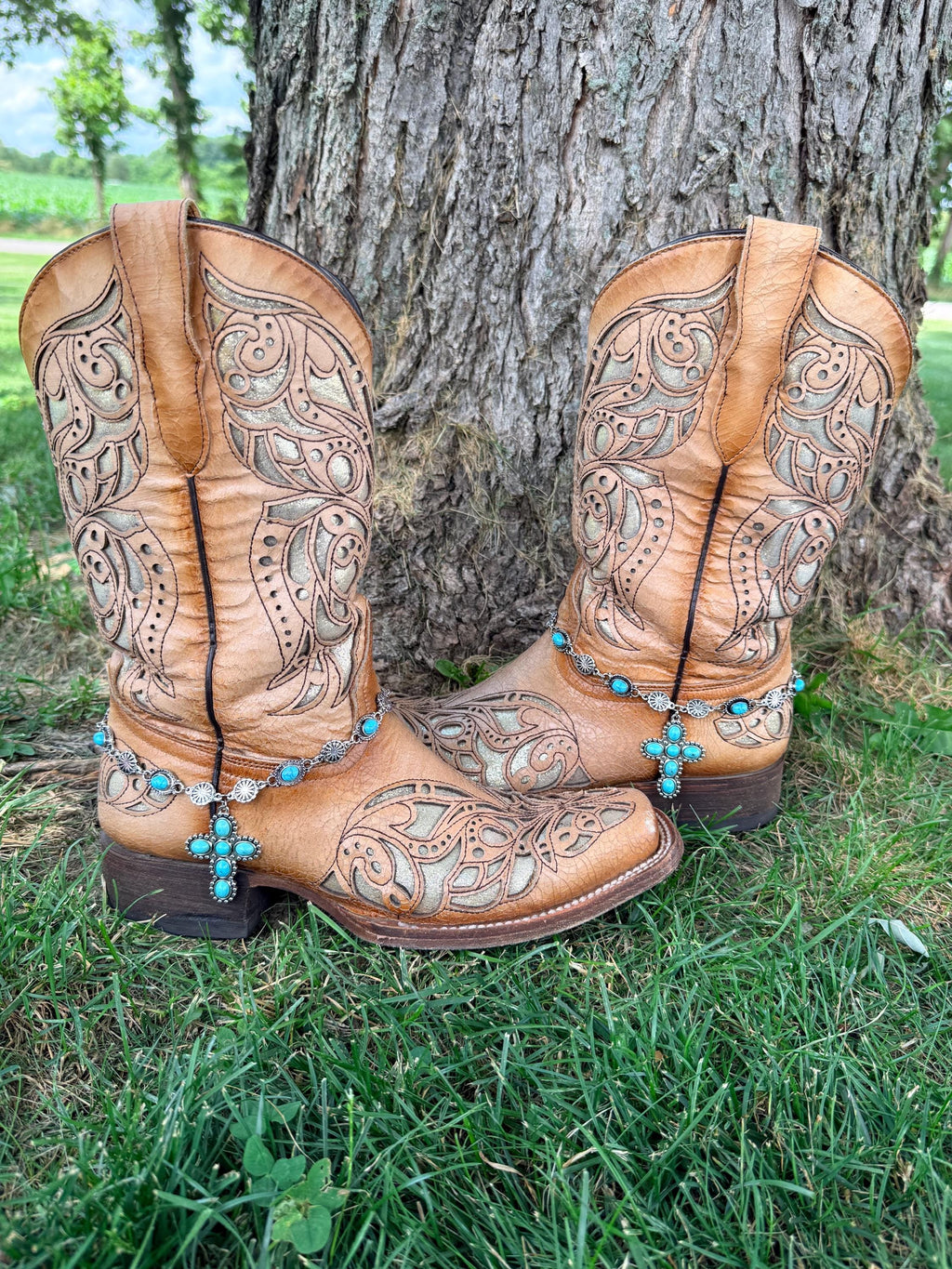 Front view of pair of tan tooled laser-cut cowboy boots against tree on grass wearing silver and turquoise concho chain boot bracelets with large six-stone turquoise cross pendant, Western boho boot jewelry set of 2