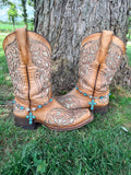 Front view of pair of tan tooled laser-cut cowboy boots against tree on grass wearing silver and turquoise concho chain boot bracelets with large six-stone turquoise cross pendant, Western boho boot jewelry set of 2