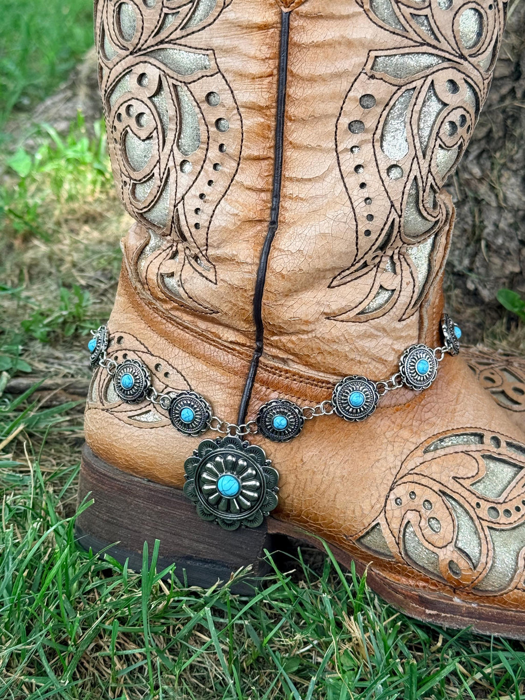 Close-up of tan tooled laser-cut cowboy boot ankle on grass with silver linked concho chain boot bracelet featuring small round turquoise-centered concho links and large sunburst-style turquoise center drop pendant