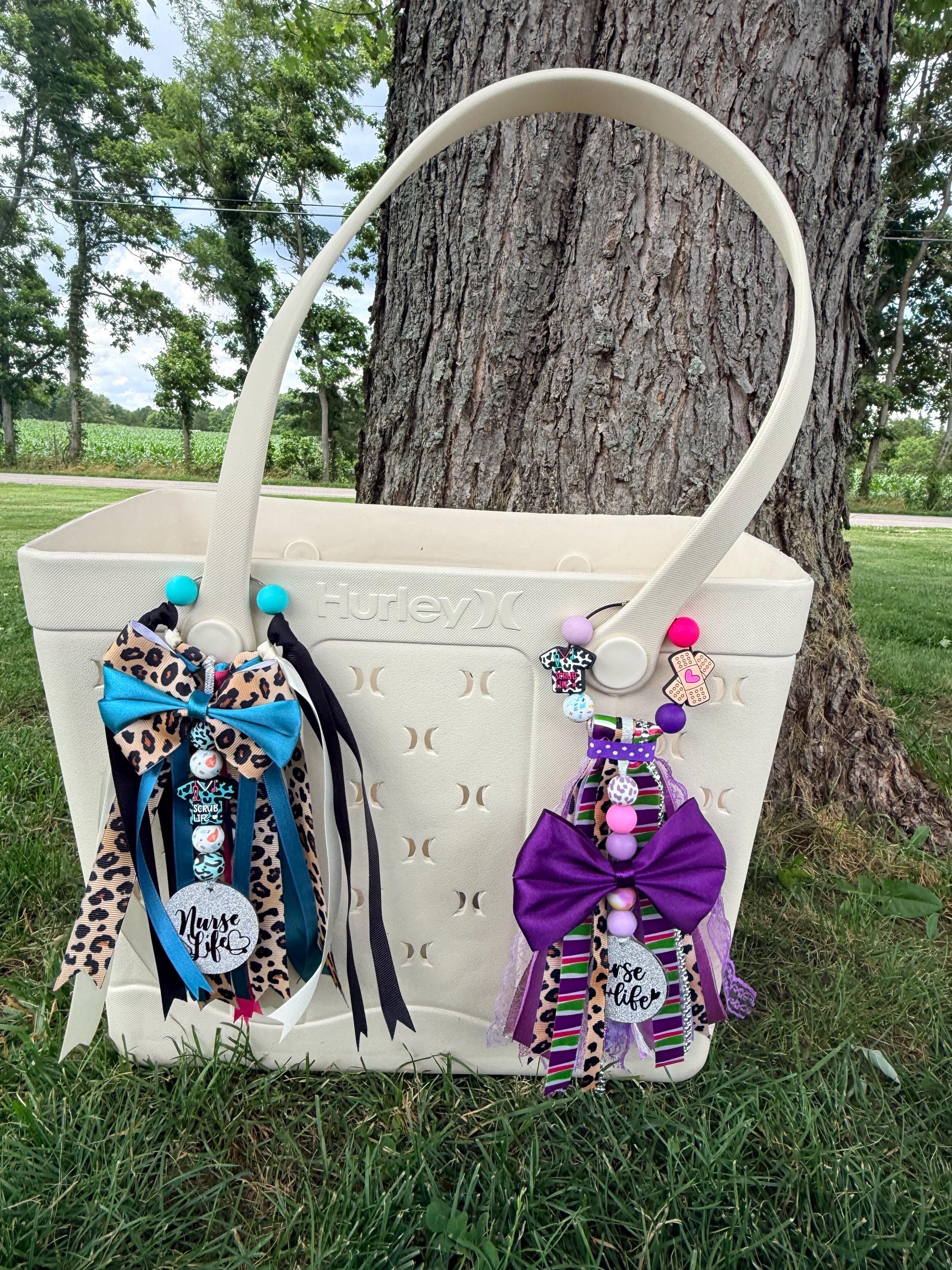 A cream-colored plastic picnic basket bag with decorative ribbons and charms, set outdoors on grass next to a large tree trunk.