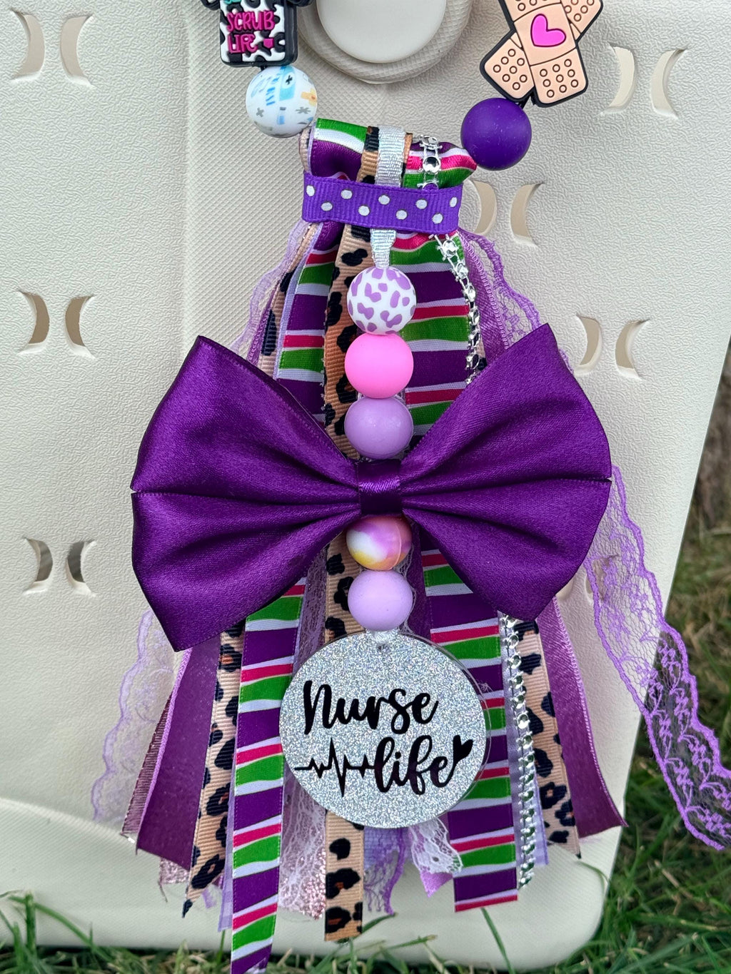 Nurse Life silicone beaded bag tassel in purple with large purple bow, multicolor striped and cheetah ribbon, scrub top and bandage charms, Nurse Life glitter charm disc, and pink and purple silicone beads clipped onto a white tote bag outdoors
