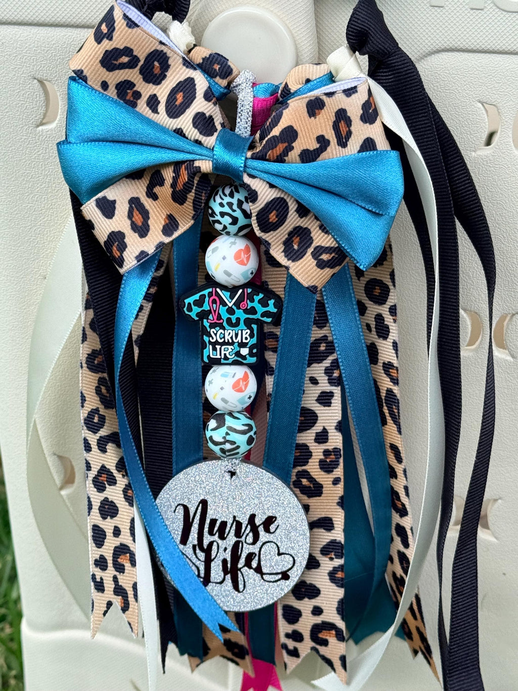 Nurse Life silicone beaded bag tassel in cheetah print with teal ribbon bow, Nurse Life glitter charm disc, scrub life bead, bandage charm, and layered ribbon tassel clipped onto a white tote bag outdoors
