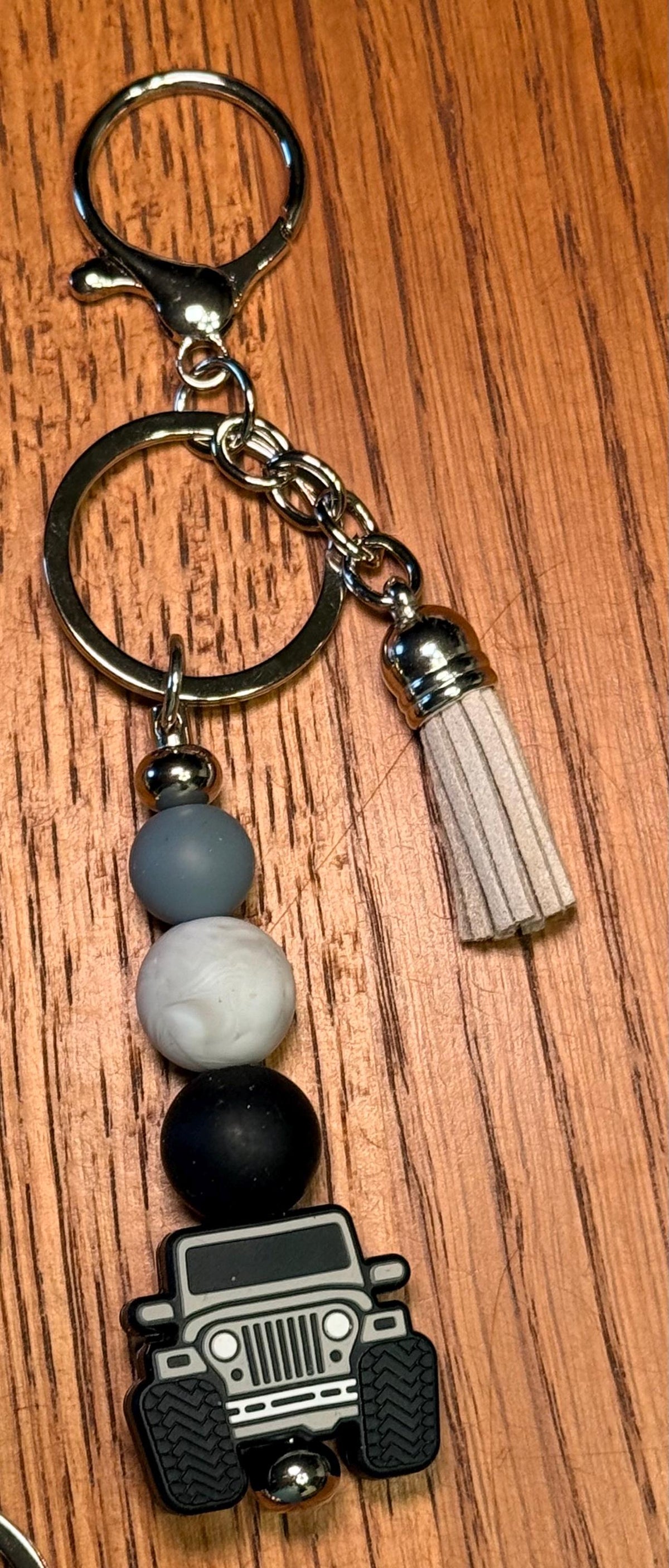 A beaded keychain with a silver-tone split ring, clasp, and tassel, featuring three matte beads in gray, white, and black, and a black off road vehicle charm at the bottom, displayed on a wooden surface.