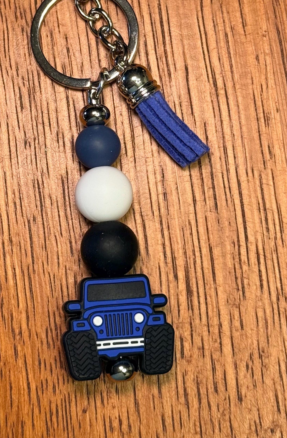 A beaded keychain featuring a blue and black off-road vehicle charm, resting on a brown wooden surface. The keychain includes a silver metal key ring, a blue tassel, and three round beads in navy blue, white, and black.