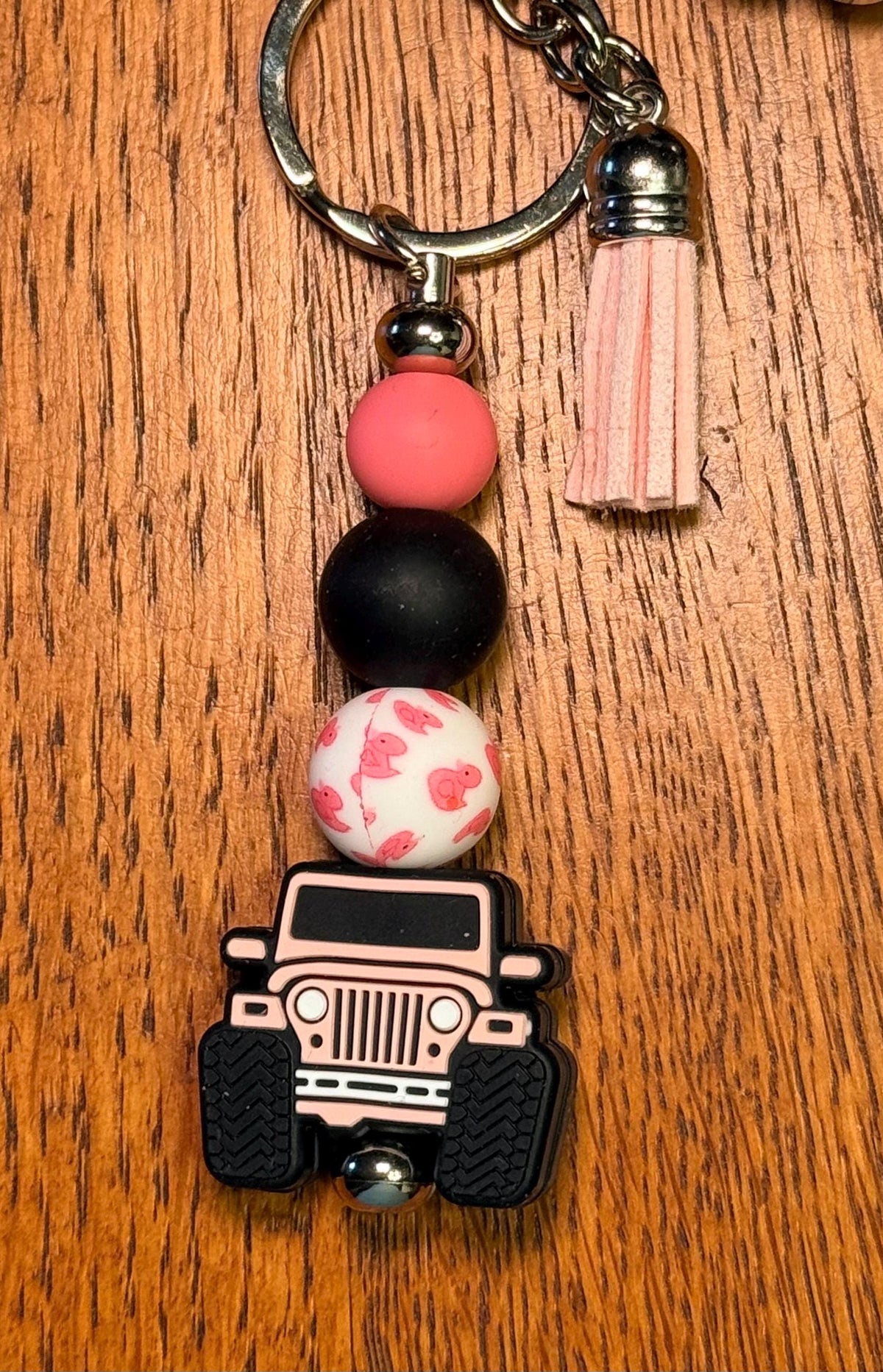 A beaded keychain with a pink and black off-road vehicle charm, resting on a wooden surface. The keychain features a silver metal ring, a pink tassel, and four decorative beads: a solid pink bead, a solid black bead, a white bead with pink floral patterns, and off-road vehicle charm with black tires.