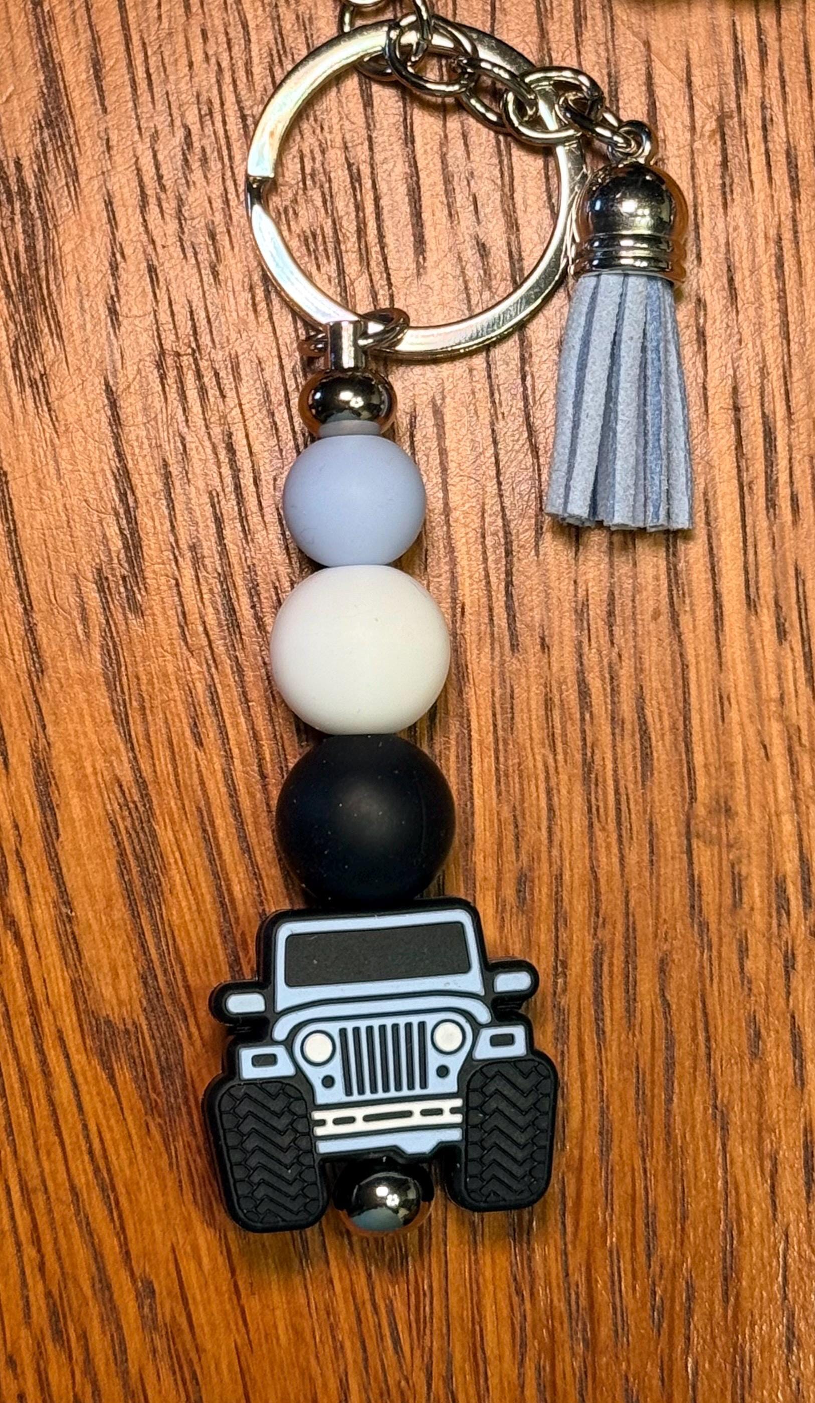 A beaded keychain featuring a black off-road vehicle charm, a gray tassel, and four matte beads in light blue, white, and black, displayed on a wooden surface.