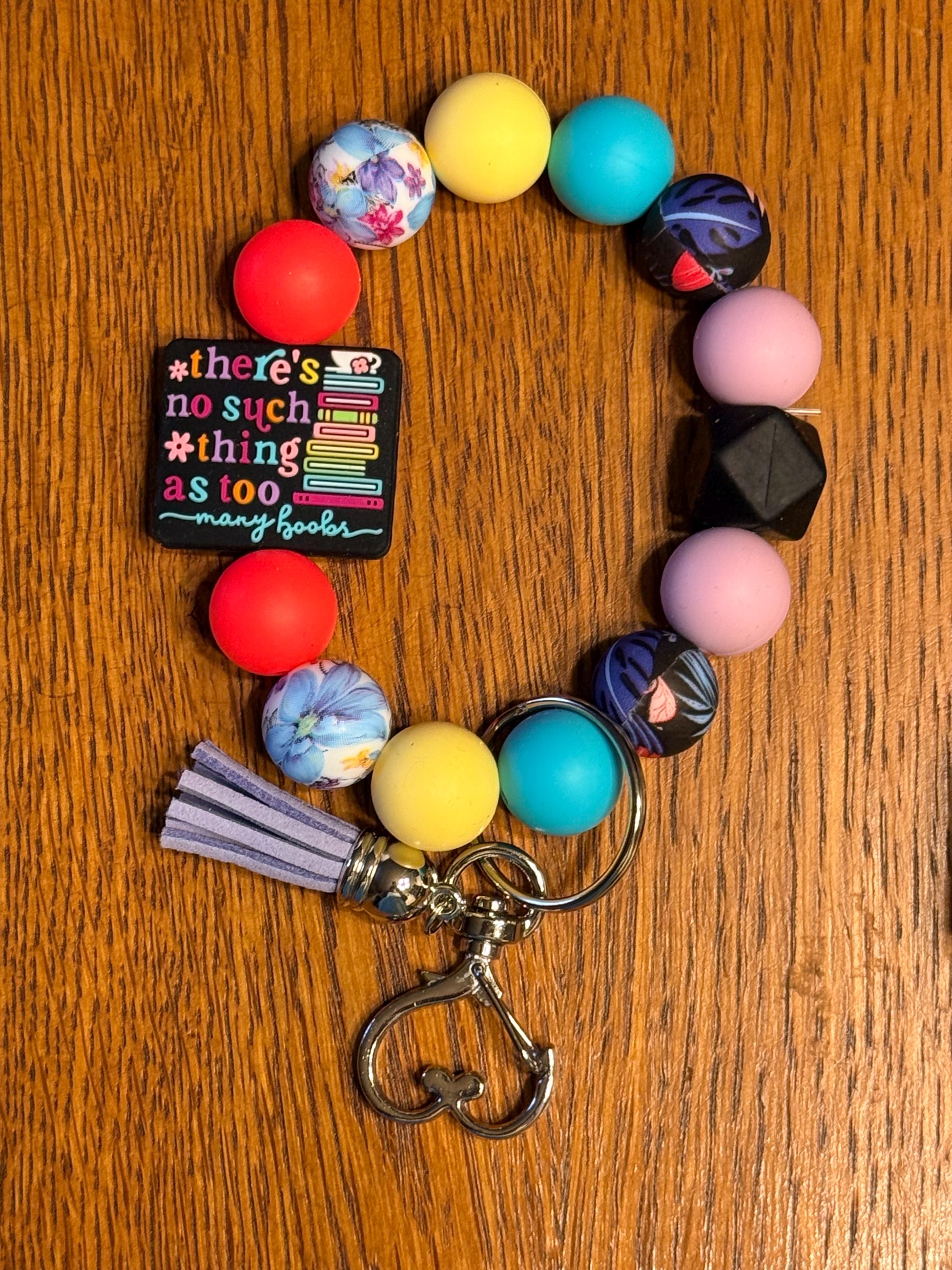 A colorful beaded keychain bracelet with a lavender tassel and silver metal clasp, displayed on a wooden surface. The beads feature various colors and patterns, including solid yellow, blue, pink, purple, and red, as well as floral and anime character designs. There's no such thing as to many books