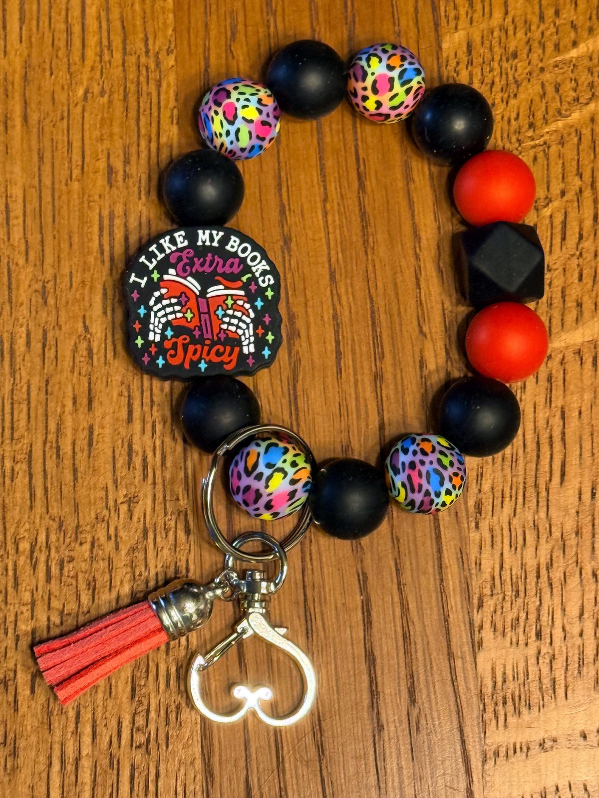 A beaded keychain bracelet on a wooden surface, featuring matte black beads, colorful leopard print beads, red and orange beads, a black guitar-shaped charm with the text 'I LIKE MY BOOKS EXTRA Spicy', and a red tassel with a silver heart clasp.