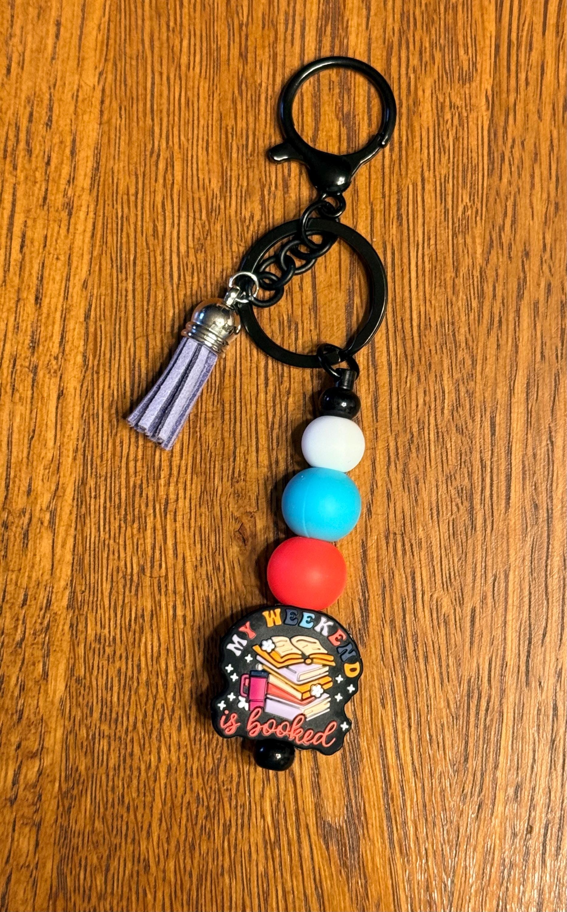 A keychain with a black metal clasp and split ring, featuring a lavender tassel charm, three round beads in white, blue, and red, and a circular enamel pin that reads 'THE WEEKEND IS BOOKED' with a book illustration, displayed on a wooden surface.