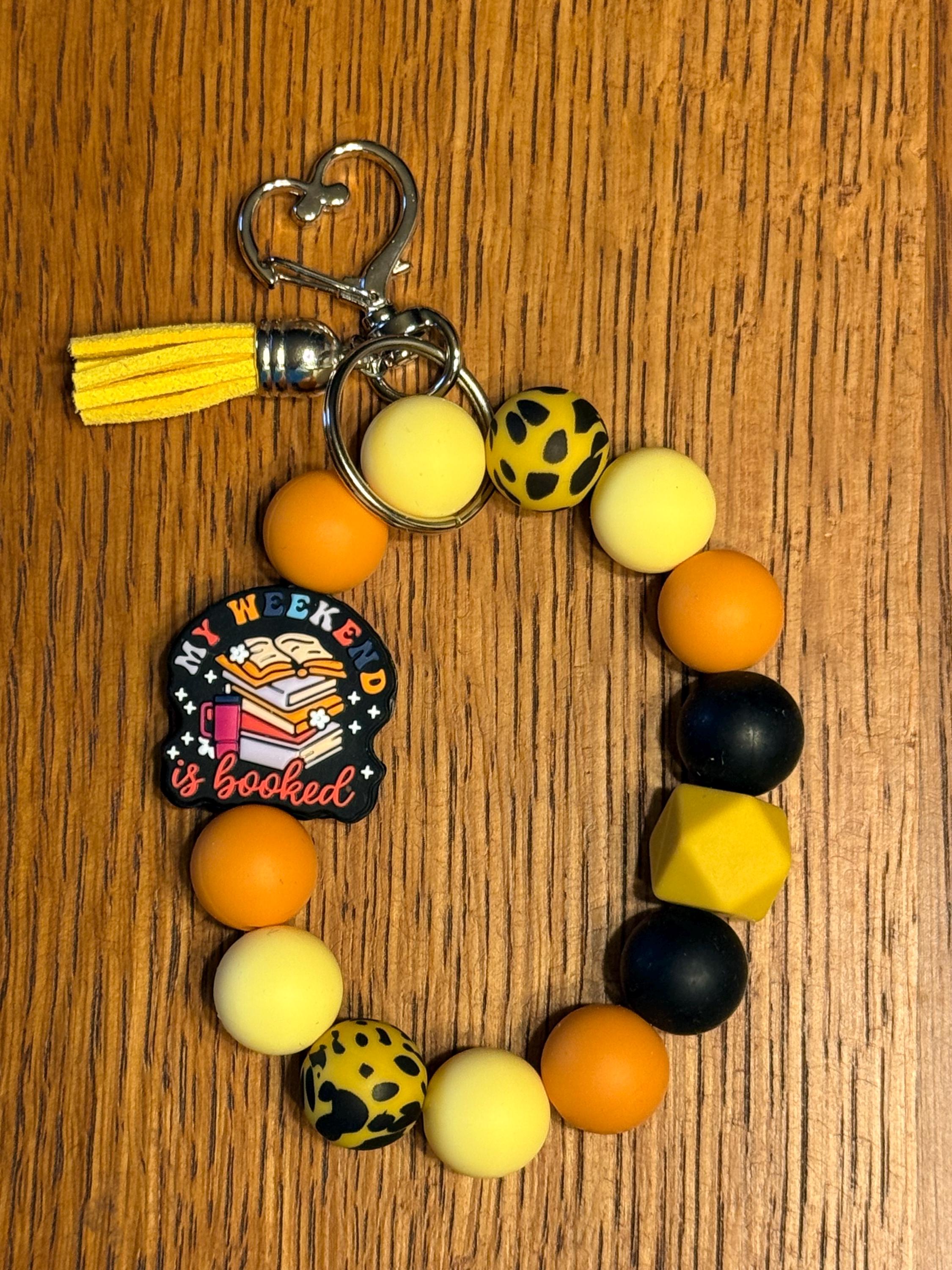 A beaded keychain with orange, pale yellow, and black leopard-print beads, a yellow tassel, silver metal rings, and a round black charm that reads 'My Weekend is Booked'. The keychain is displayed on a wooden surface.