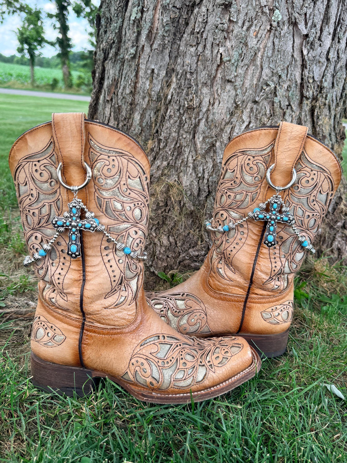 Front view of pair of tan laser-cut scrollwork cowboy boots on grass against tree wearing silver ball chain and turquoise bead boot bracelet leis with large ornate silver cross pendant set with turquoise cabochon stones, Western boot bling set of 2