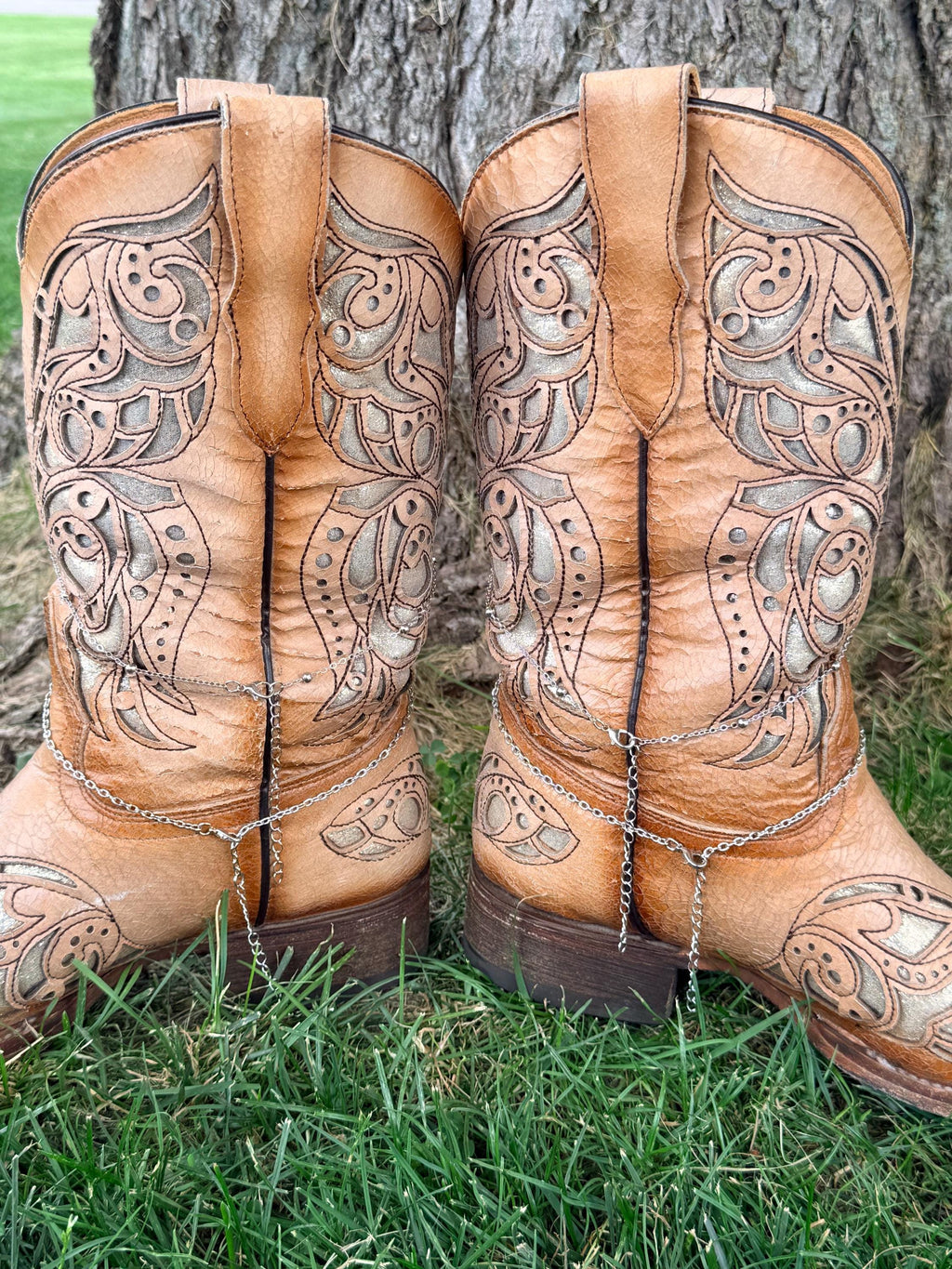 Back heel view of pair of tan tooled laser-cut cowboy boots against tree wearing silver multi-chain boot bracelets with turquoise concho charms, Western boho style
