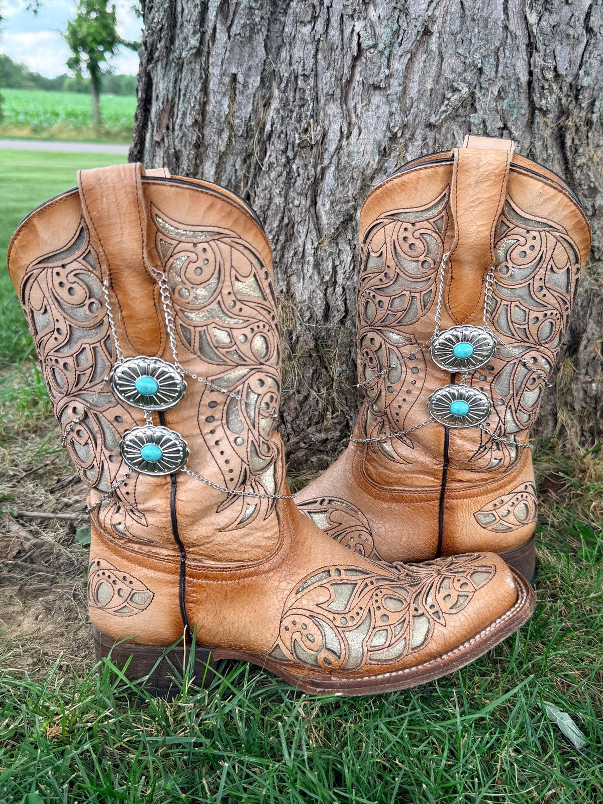 Front view of pair of tan tooled laser-cut cowboy boots against tree with silver multi-chain boot bracelets featuring two stacked turquoise oval concho charms, Western boho boot jewelry set of 2