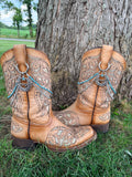 Front view of pair of tan tooled laser-cut cowboy boots against tree on grass wearing two-strand turquoise bead and silver chain lei boot bracelets with large silver horseshoe cowgirl charm, set of 2
