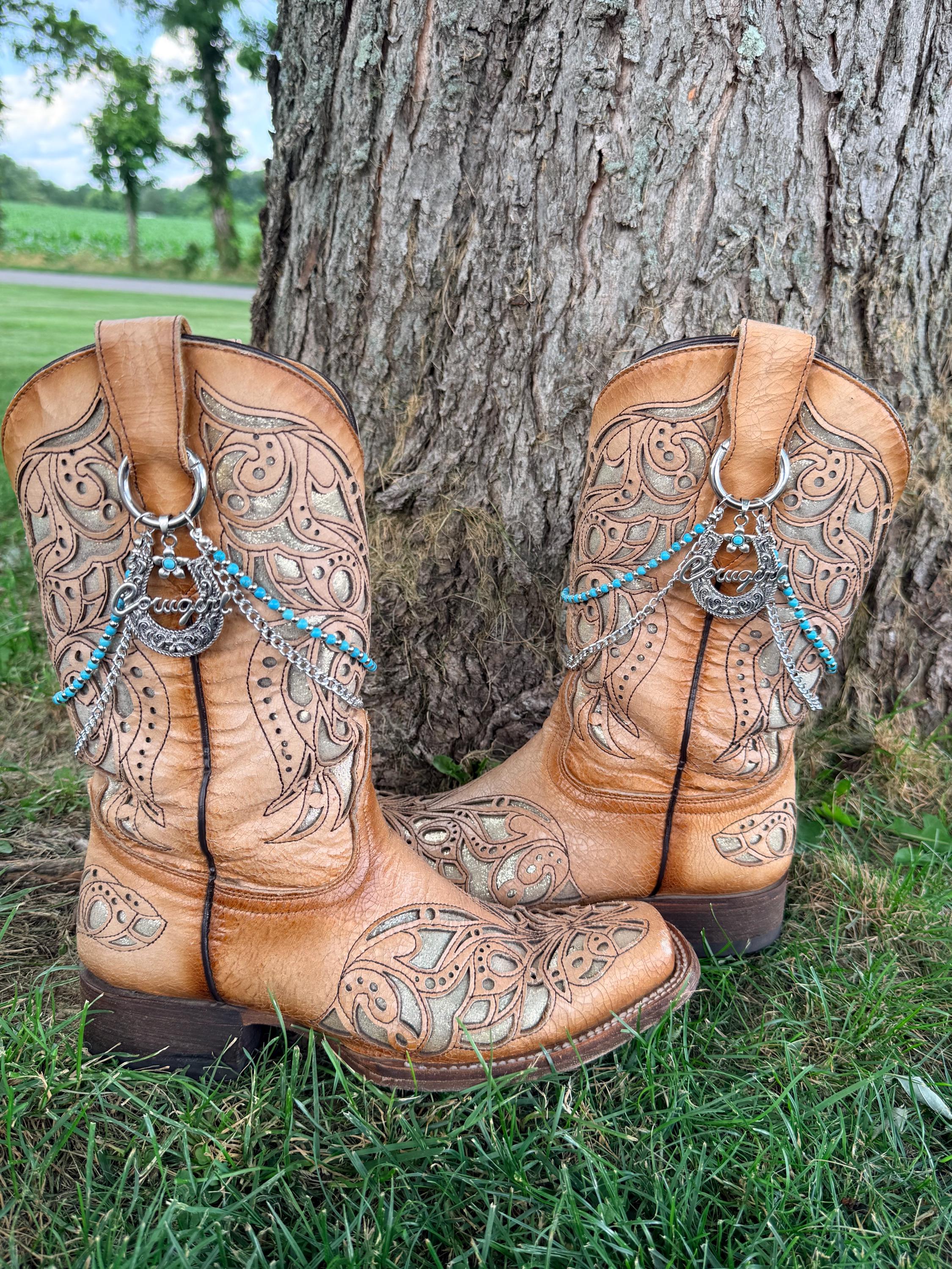 Front view of pair of tan tooled laser-cut cowboy boots against tree on grass wearing two-strand turquoise bead and silver chain lei boot bracelets with large silver horseshoe cowgirl charm, set of 2
