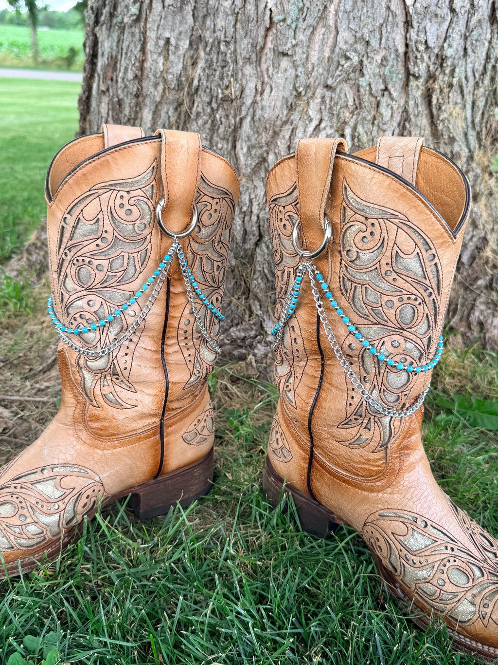 Back view of pair of tan tooled laser-cut cowboy boots against tree showing two-strand turquoise bead and silver chain lei boot bracelets clipped through boot pull loop rings, Western boho style set of 2