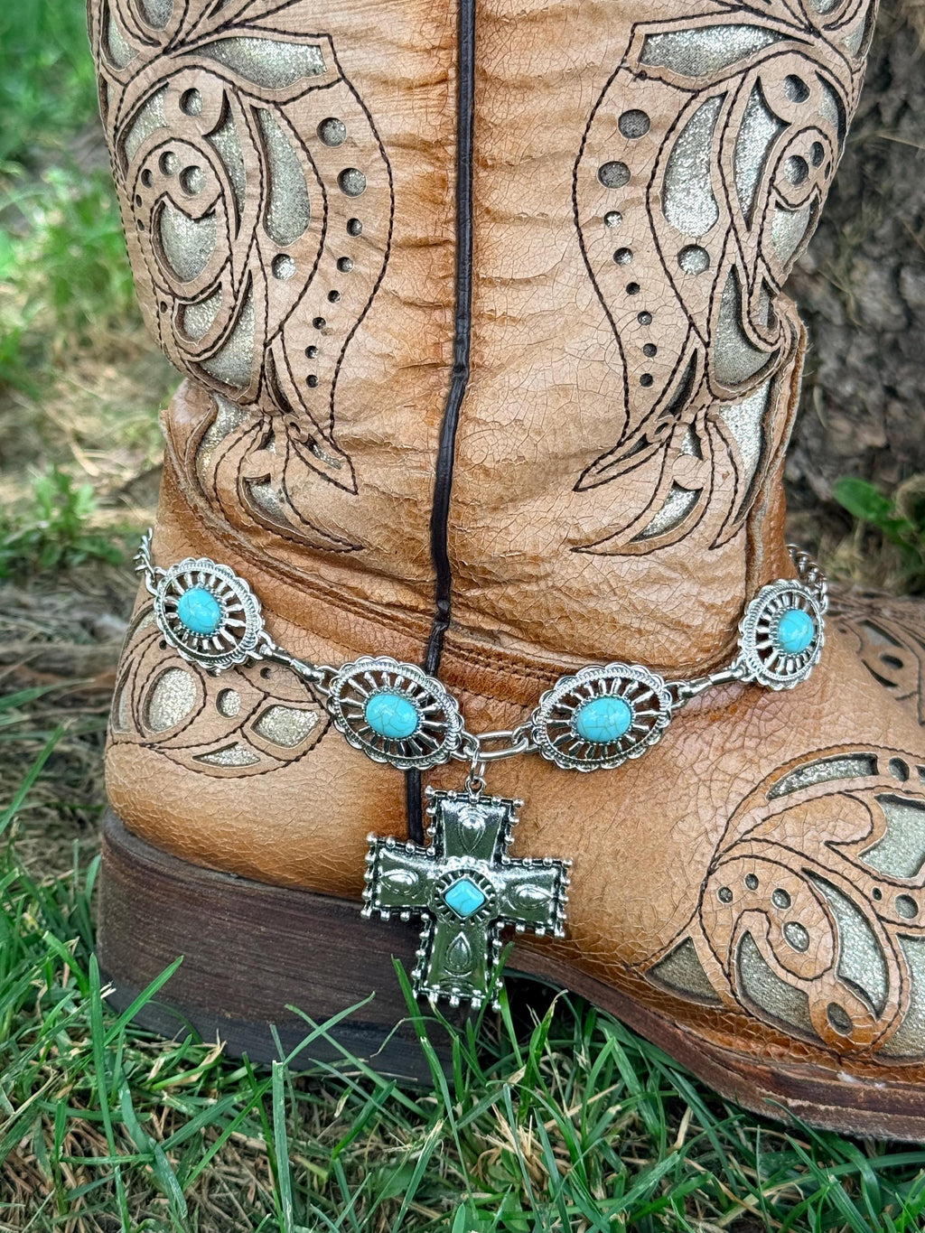 Close-up of tan tooled laser-cut cowboy boot ankle on grass with large oval turquoise concho chain boot bracelet and large silver engraved cross pendant with single turquoise diamond-shaped center stone