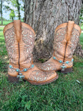 Front view of pair of tan tooled laser-cut cowboy boots against tree on grass wearing large oval turquoise concho chain boot bracelets with large silver engraved cross pendant and turquoise center stone, Western boho boot jewelry set of 2