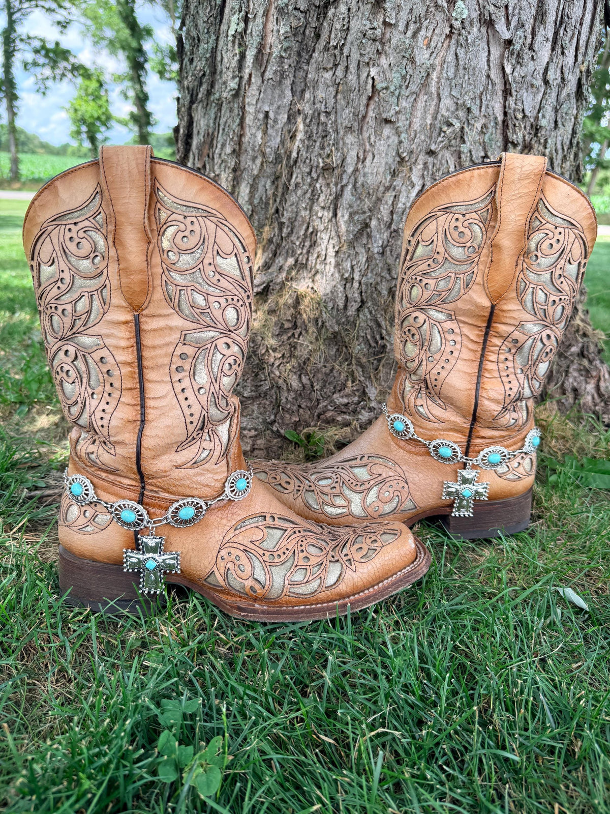 Front view of pair of tan tooled laser-cut cowboy boots against tree on grass wearing large oval turquoise concho chain boot bracelets with large silver engraved cross pendant and turquoise center stone, Western boho boot jewelry set of 2