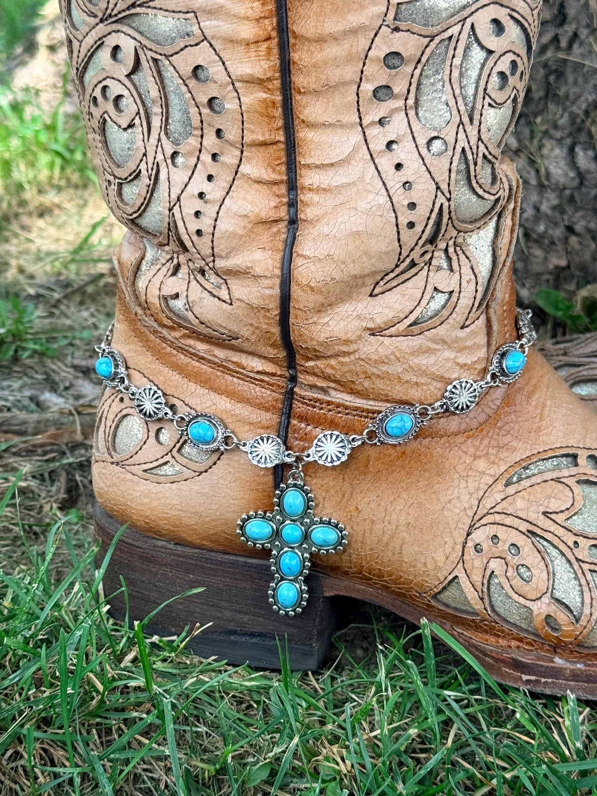 Close-up of tan tooled laser-cut cowboy boot ankle on grass with silver chain boot bracelet featuring alternating oval turquoise cabochon and silver sunburst concho links and large six-stone turquoise cross pendant