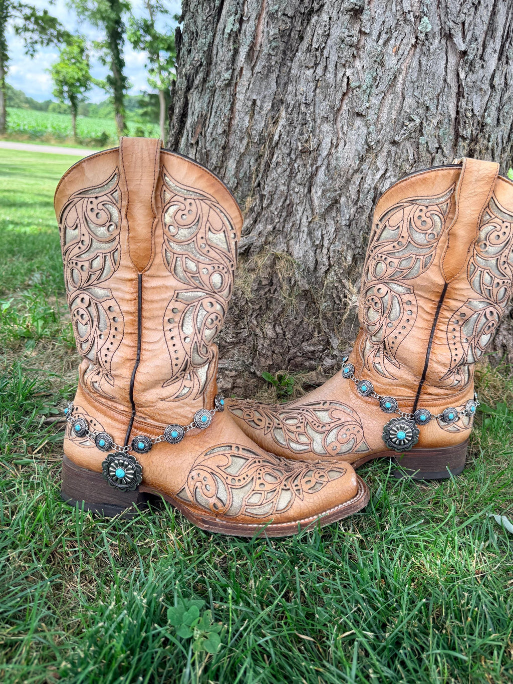 Front view of pair of tan tooled laser-cut cowboy boots against tree on grass wearing silver linked concho chain boot bracelets with large sunburst-style turquoise center drop pendant, Western boho boot jewelry set of 2