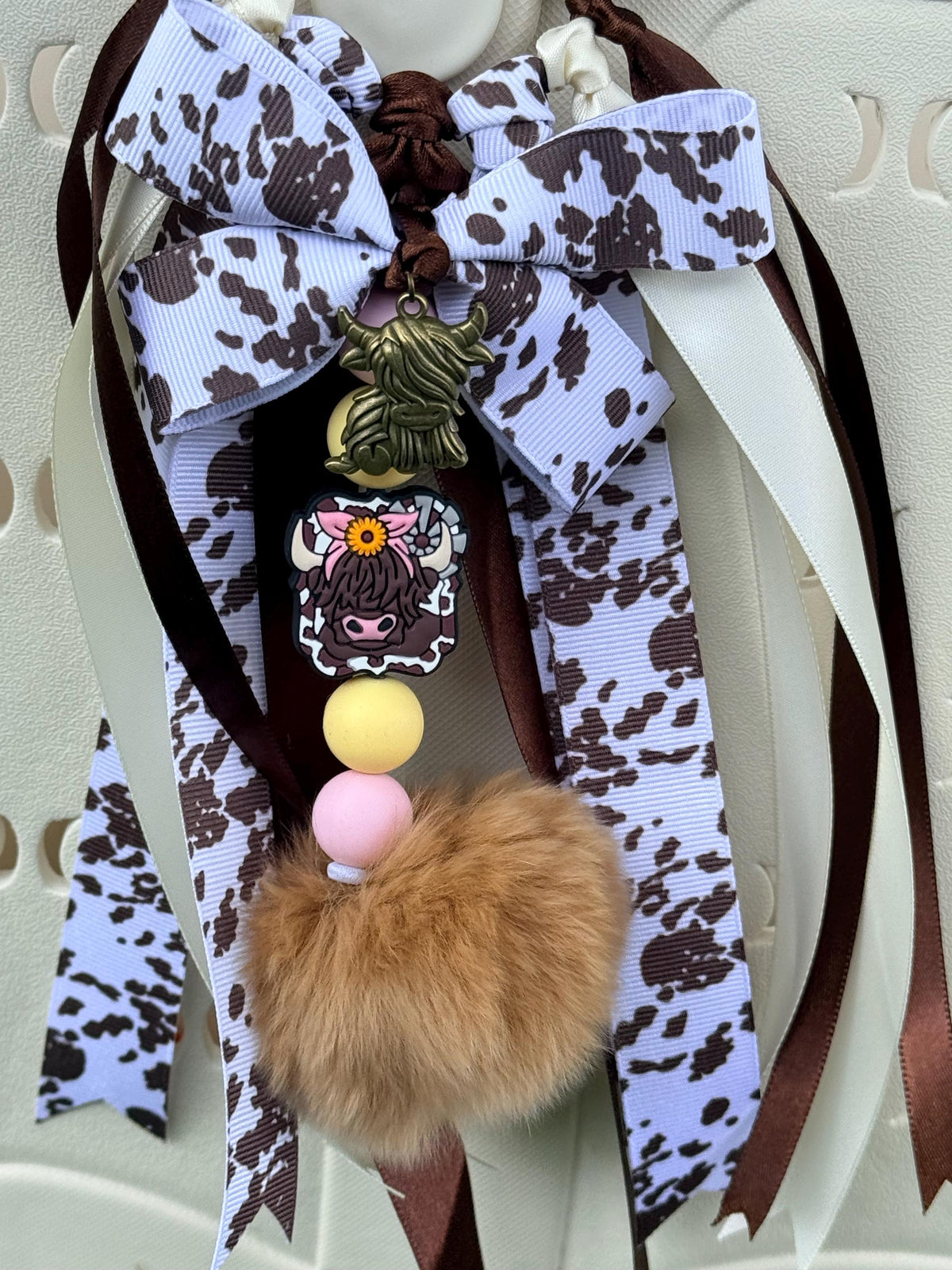 Highland Cow silicone beaded bag tassel with cow print ribbon bow, highland cow charm, yellow and pink beads, and faux fur pom pom, shown clipped onto a white tote bag
