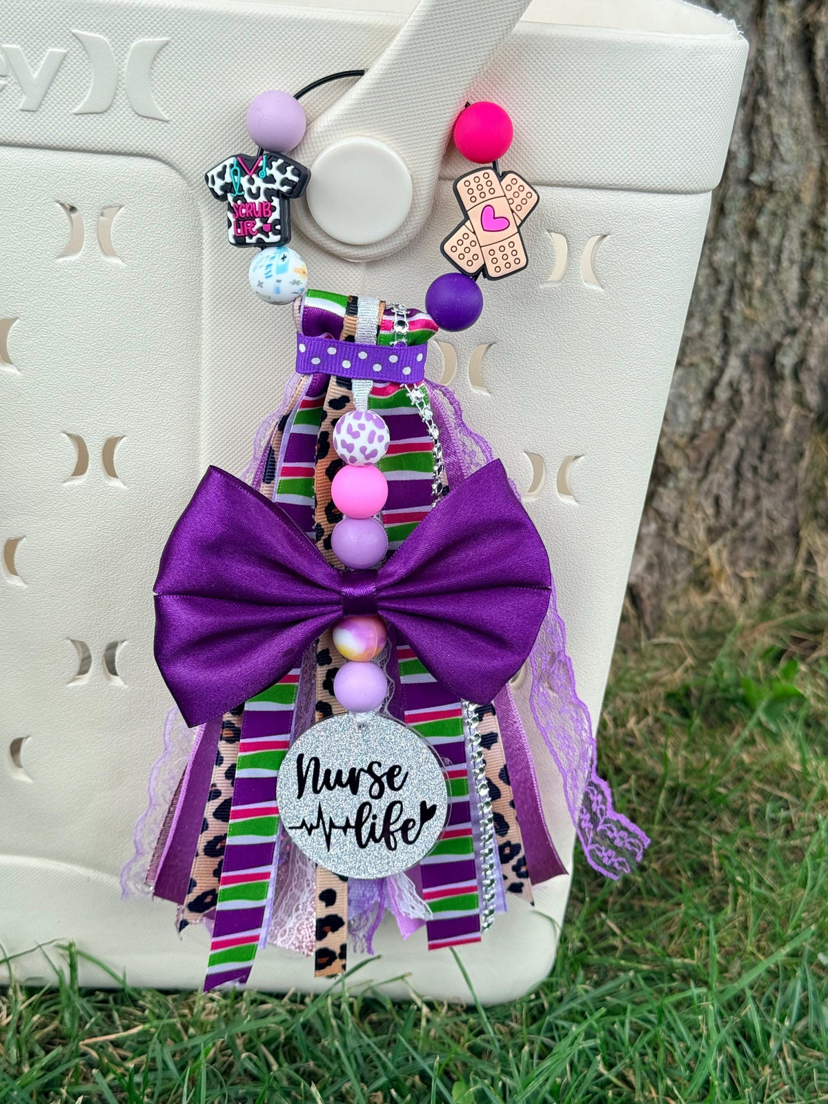 Nurse Life silicone beaded bag tassel in purple with large purple bow, multicolor striped and cheetah ribbon, scrub top and bandage charms, Nurse Life glitter charm disc, and pink and purple silicone beads clipped onto a white tote bag outdoors

