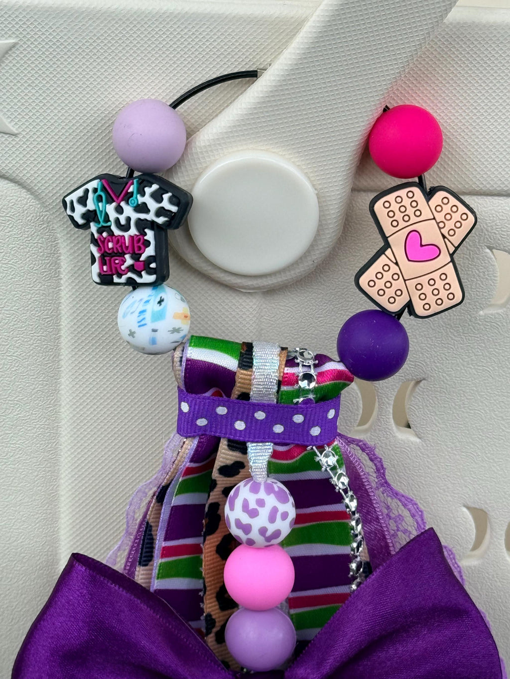 Nurse Life silicone beaded bag tassel in purple with large purple bow, multicolor striped and cheetah ribbon, scrub top and bandage charms, Nurse Life glitter charm disc, and pink and purple silicone beads clipped onto a white tote bag outdoors
