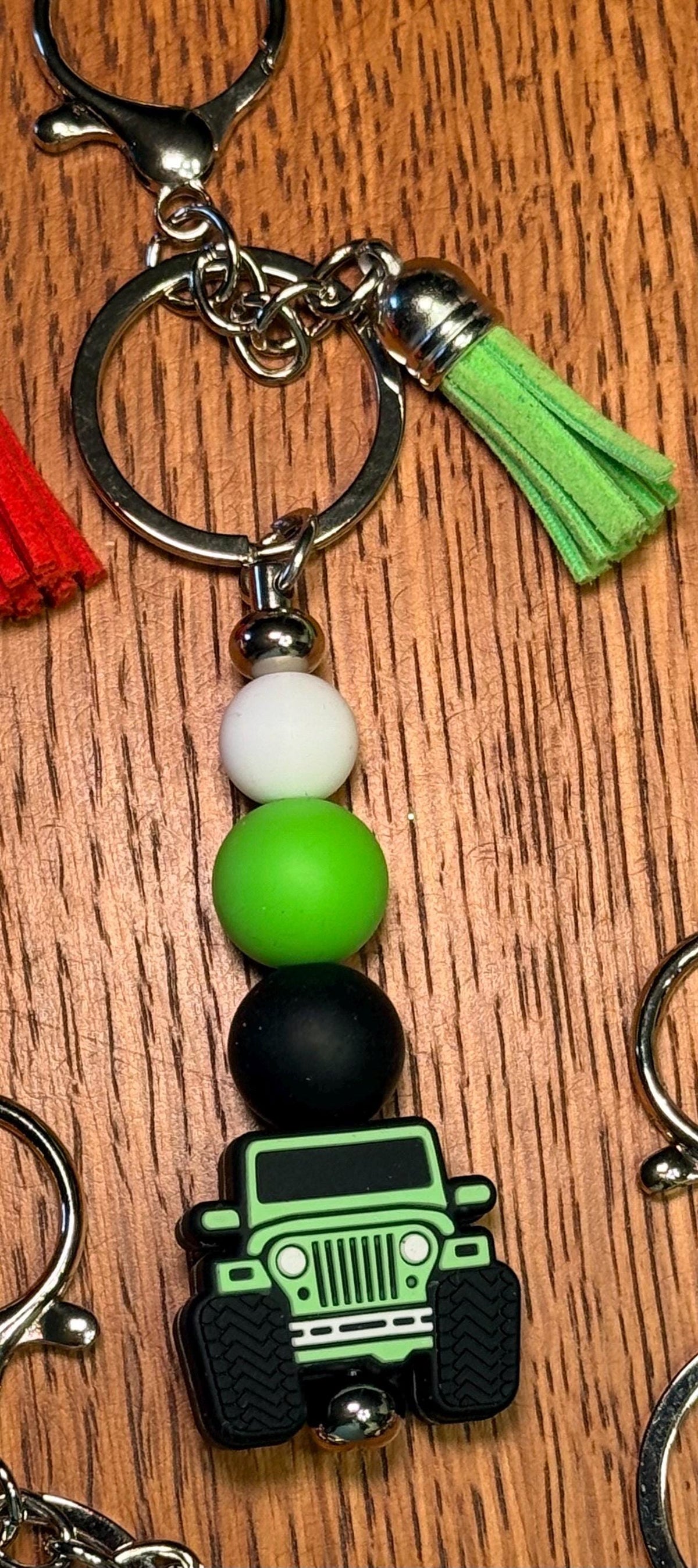 A close-up of a keychain featuring a green and black off-road vehicle charm, matte beads in white, green, and black, a green tassel, and a silver key ring, displayed on a wooden surface.